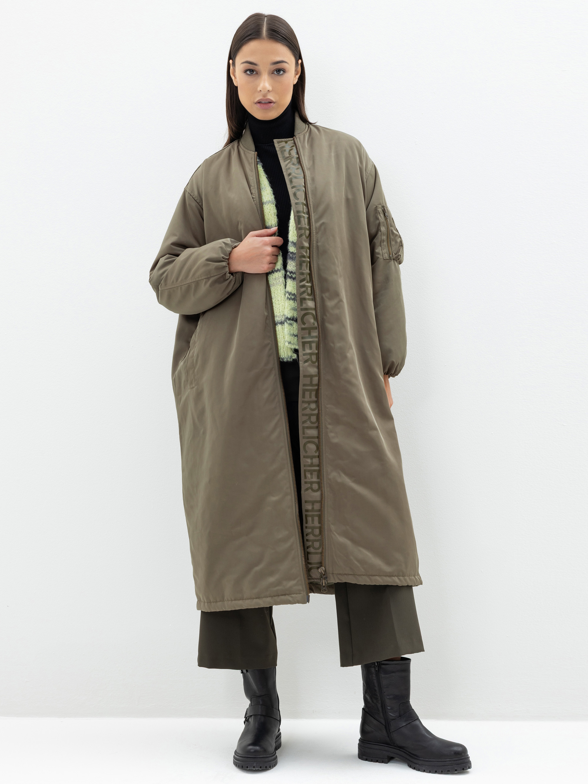 Eine Frau steht vor einem weißen Hintergrund und trägt einen langen, olivgrünen Mantel über einer dunklen, weit geschnittenen Hose, schwarze Stiefel und einen schwarzen Rollkragenpullover. Sie blickt in die Kamera und hält den Mantel mit einer Hand l