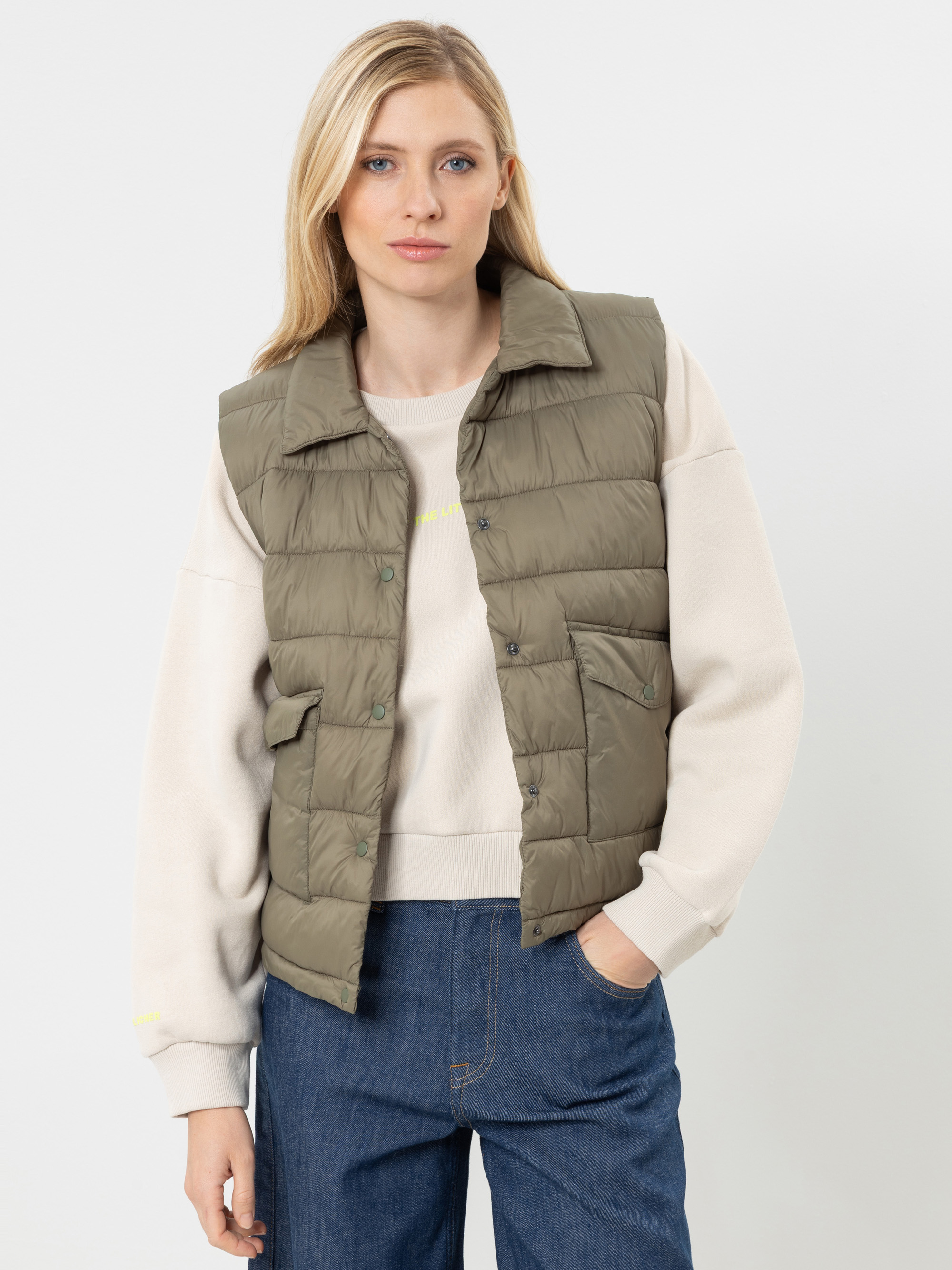Vor einem schlichten Hintergrund steht eine Frau, die einen beigen Pullover, eine olivgrüne Daunenweste und blaue Jeans trägt. Sie hat langes blondes Haar und einen ausdruckslosen Gesichtsausdruck; eine Hand steckt in ihrer Hosentasche.