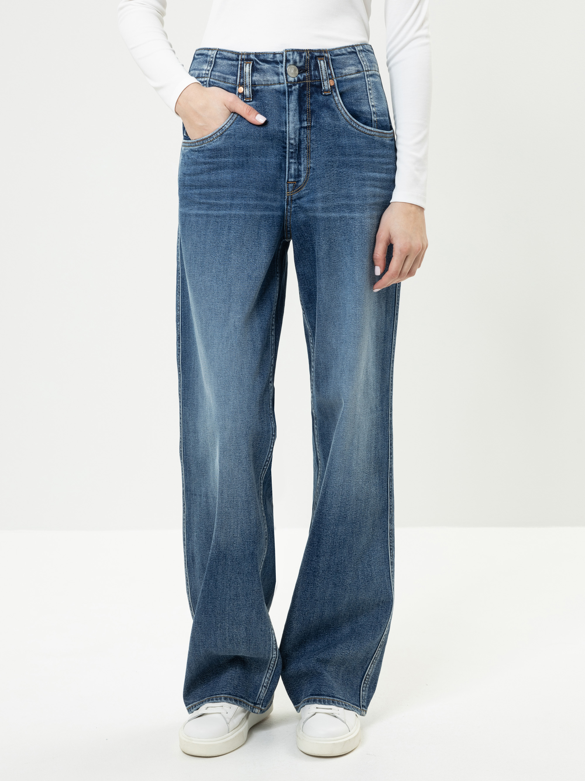 Eine Person, die eine blaue Jeans mit weitem Bein, ein weißes langärmeliges Oberteil und weiße Turnschuhe trägt, steht vor einem einfarbig weißen Hintergrund und hat eine Hand in der Tasche.