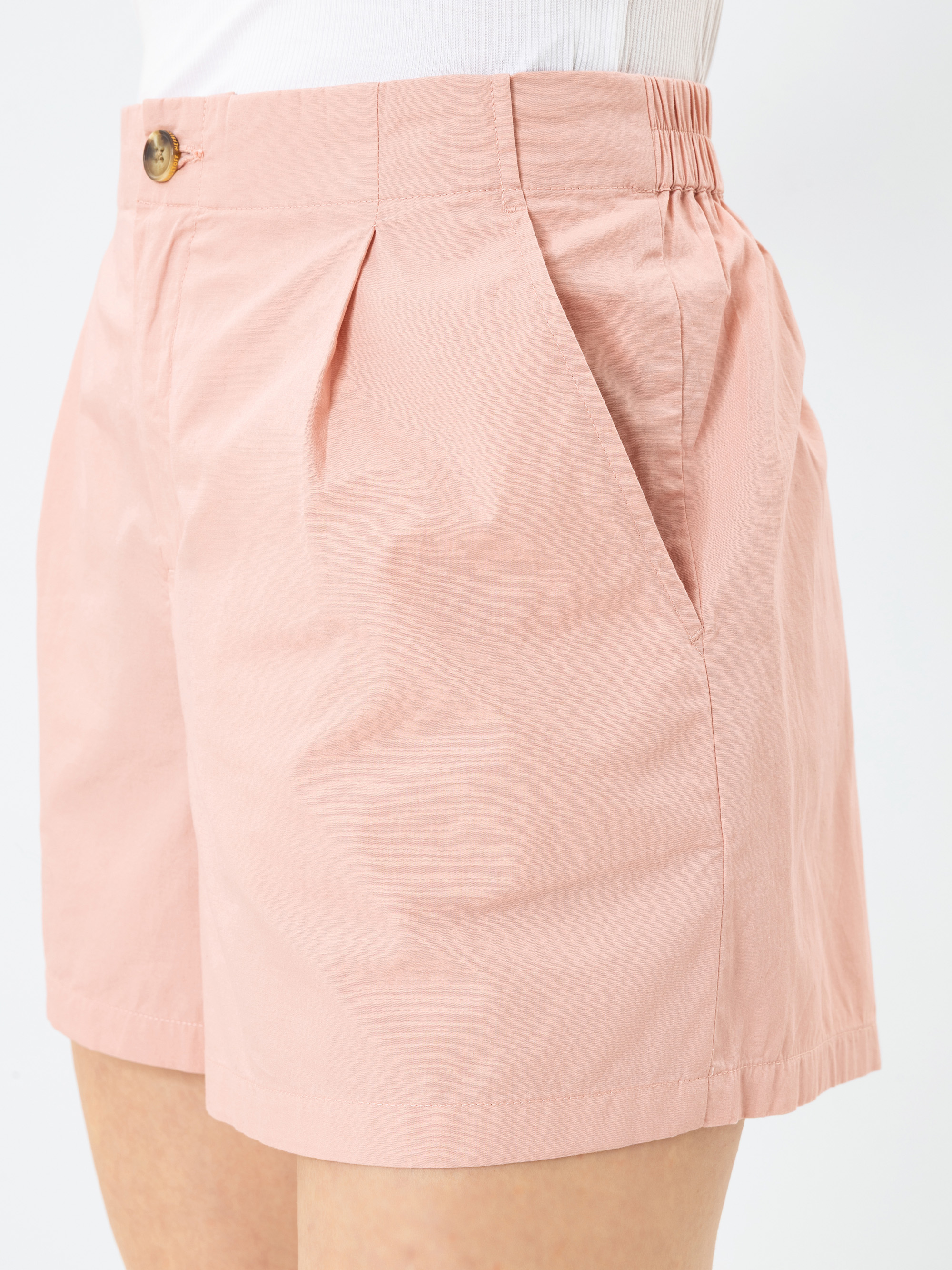 Person in hellrosa Shorts mit hoher Taille, Bundfalten, Seitentaschen, einem elastischen Bund auf der Rückseite und einem Knopfverschluss. Die Shorts werden mit einem weißen Oberteil zum Hineinstecken kombiniert.