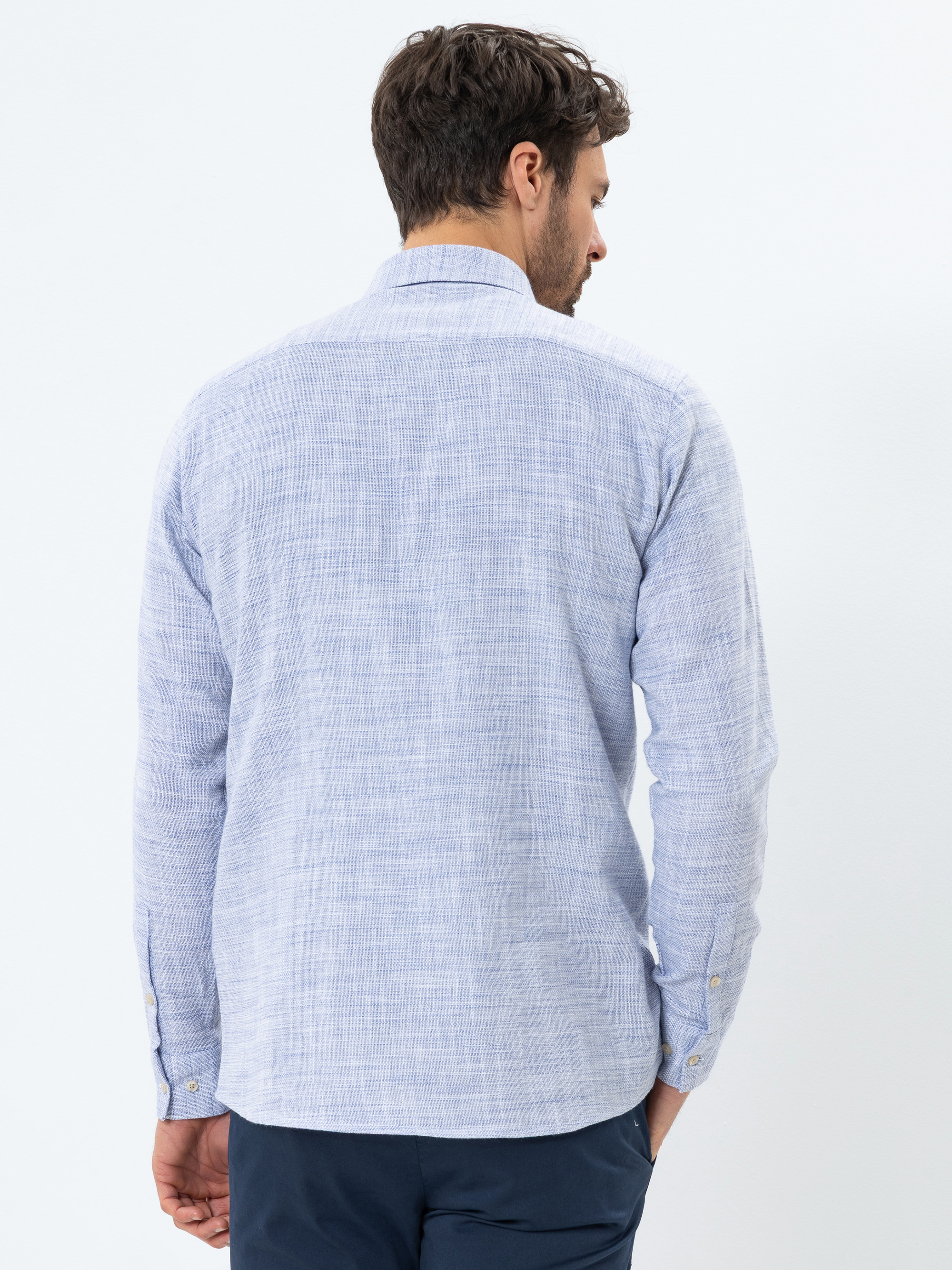 Ein Mann steht mit dem Rücken zur Kamera und trägt ein hellblaues, langärmeliges, strukturiertes Button-up-Hemd und eine dunkle Hose vor einem schlichten weißen Hintergrund.