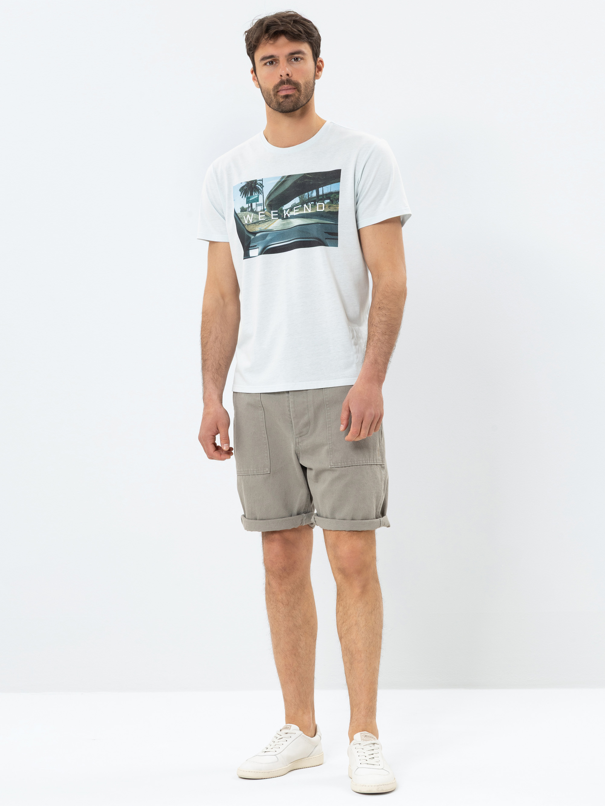 Ein Mann steht vor einem schlichten weißen Hintergrund und trägt ein weißes grafisches T-Shirt mit der Aufschrift WEEKEND, beigefarbene knielange Shorts und weiße Turnschuhe. Er hat kurzes dunkles Haar und einen Bart und einen neutralen Gesichtsausdru