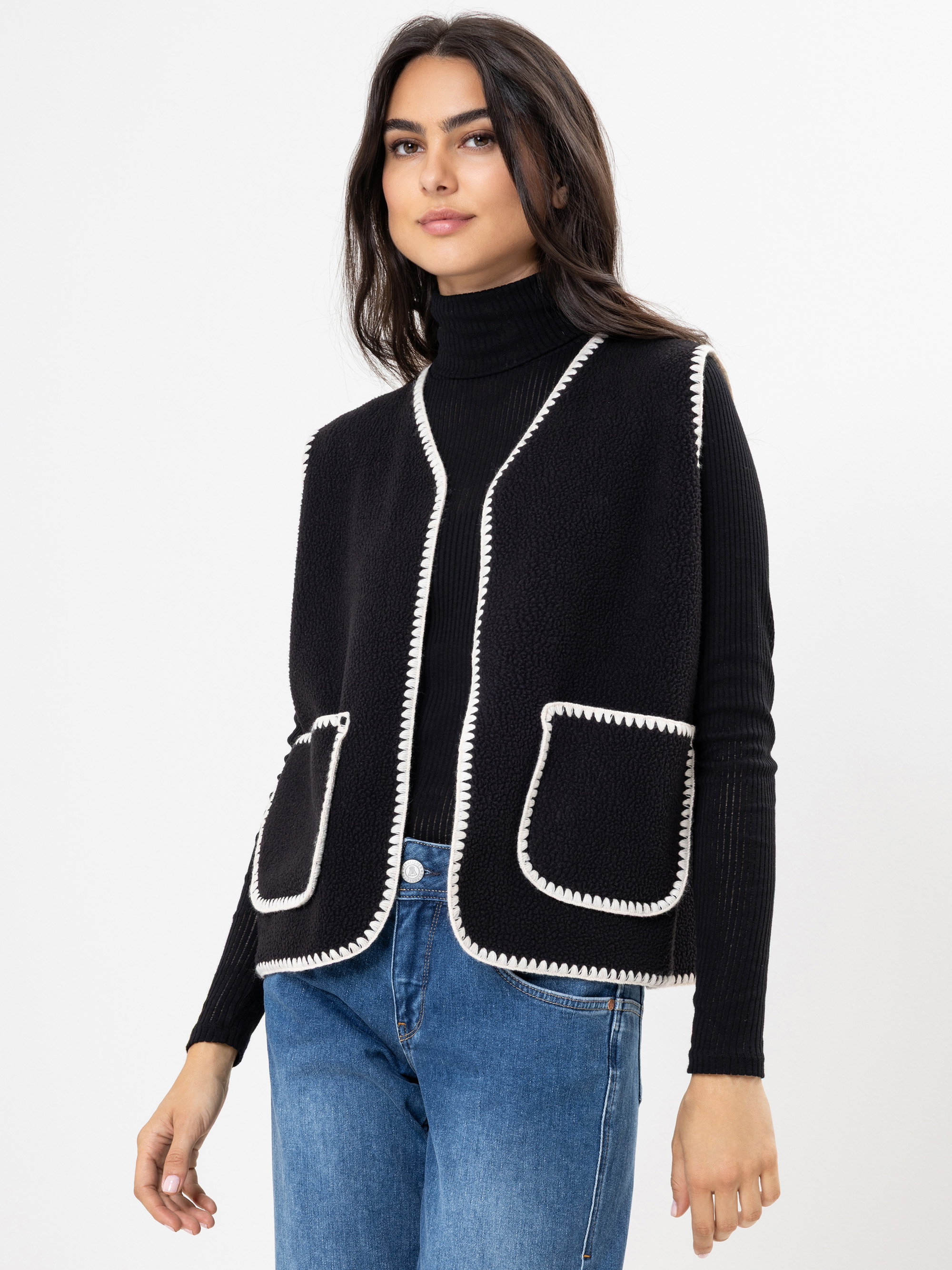 Eine Frau mit langen dunklen Haaren trägt eine schwarze ärmellose Weste mit weißem Saum, einen schwarzen Rollkragenpullover und blaue Jeans, die vor einem schlichten weißen Hintergrund stehen.
