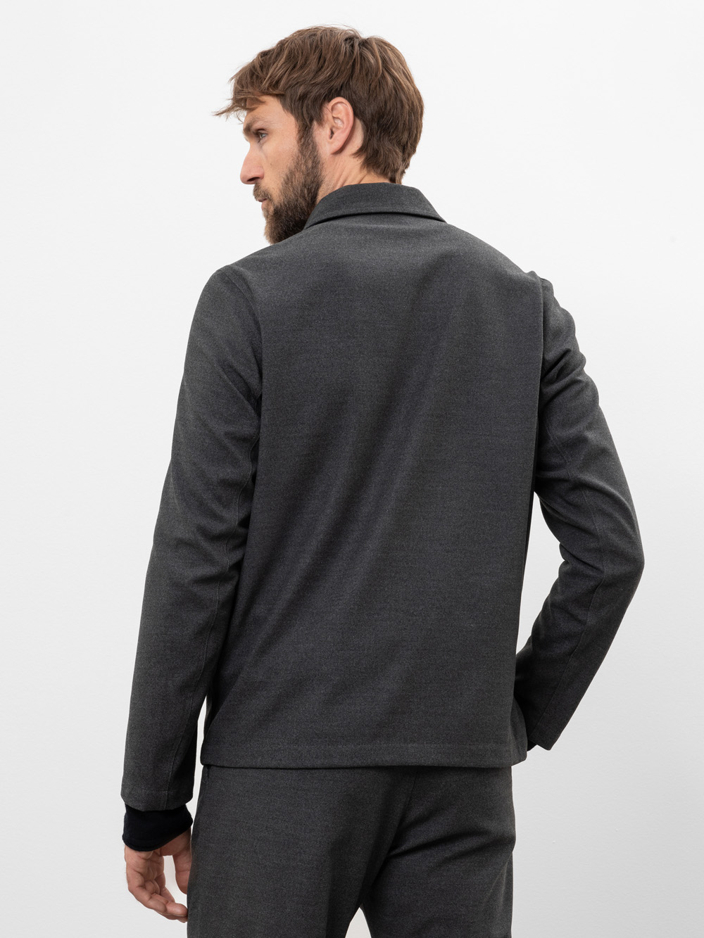 Ein Mann mit hellbraunem Haar und Bart steht mit dem Rücken zur Kamera, trägt eine dunkelgraue Anzugsjacke und eine dazu passende Hose vor einem schlichten weißen Hintergrund.