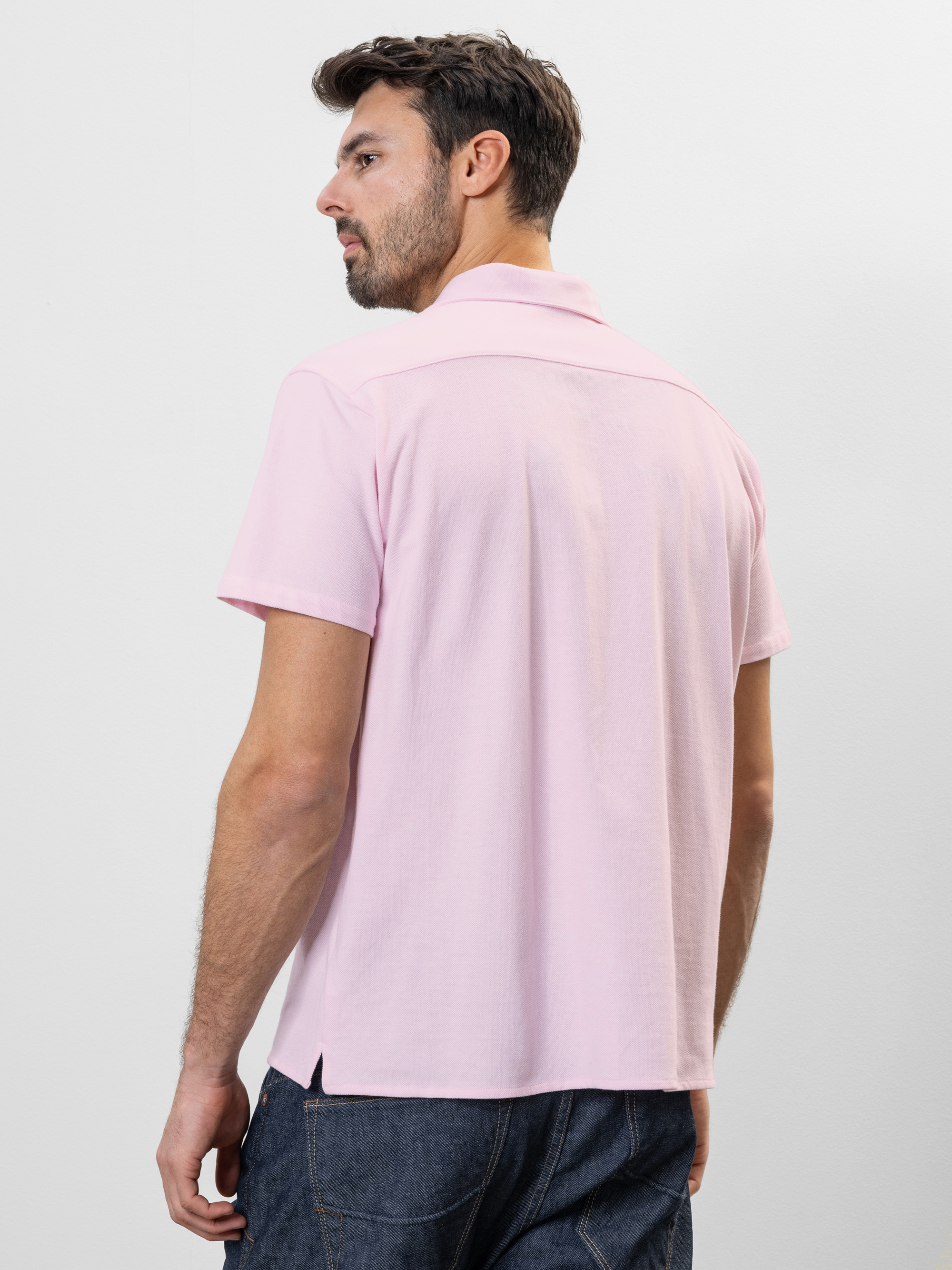Ein Mann mit kurzen braunen Haaren und Bart trägt ein hellrosa, kurzärmeliges Polohemd und dunkle Jeans. Er steht vor einem schlichten weißen Hintergrund und blickt von der Kamera weg.