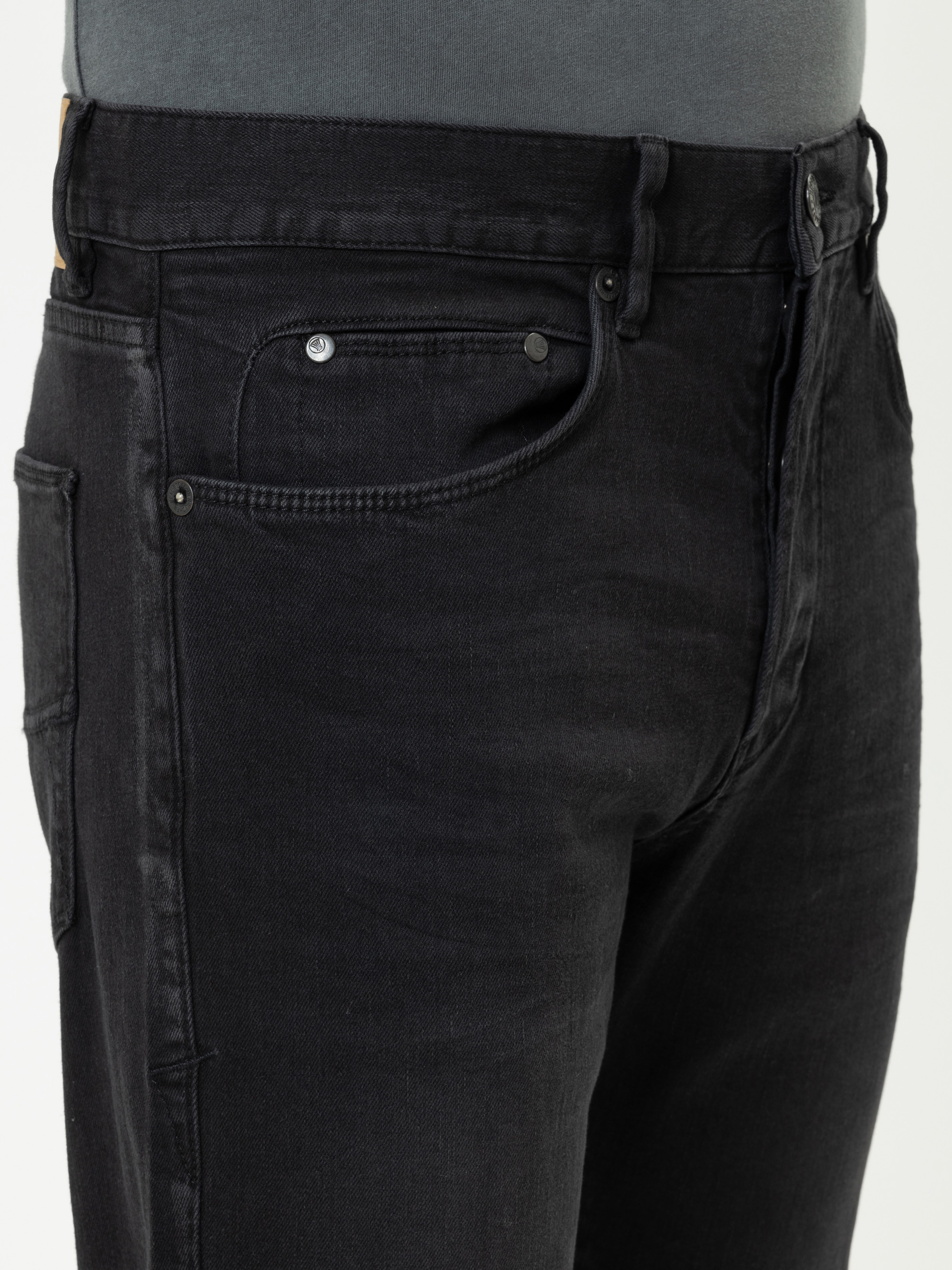 Eine Person, die eine schwarze Jeans und ein graues Hemd trägt, wird von der Taille bis zur Mitte des Oberschenkels gezeigt, wobei der Schwerpunkt auf der vorderen Tasche und dem Bund der Jeans liegt.