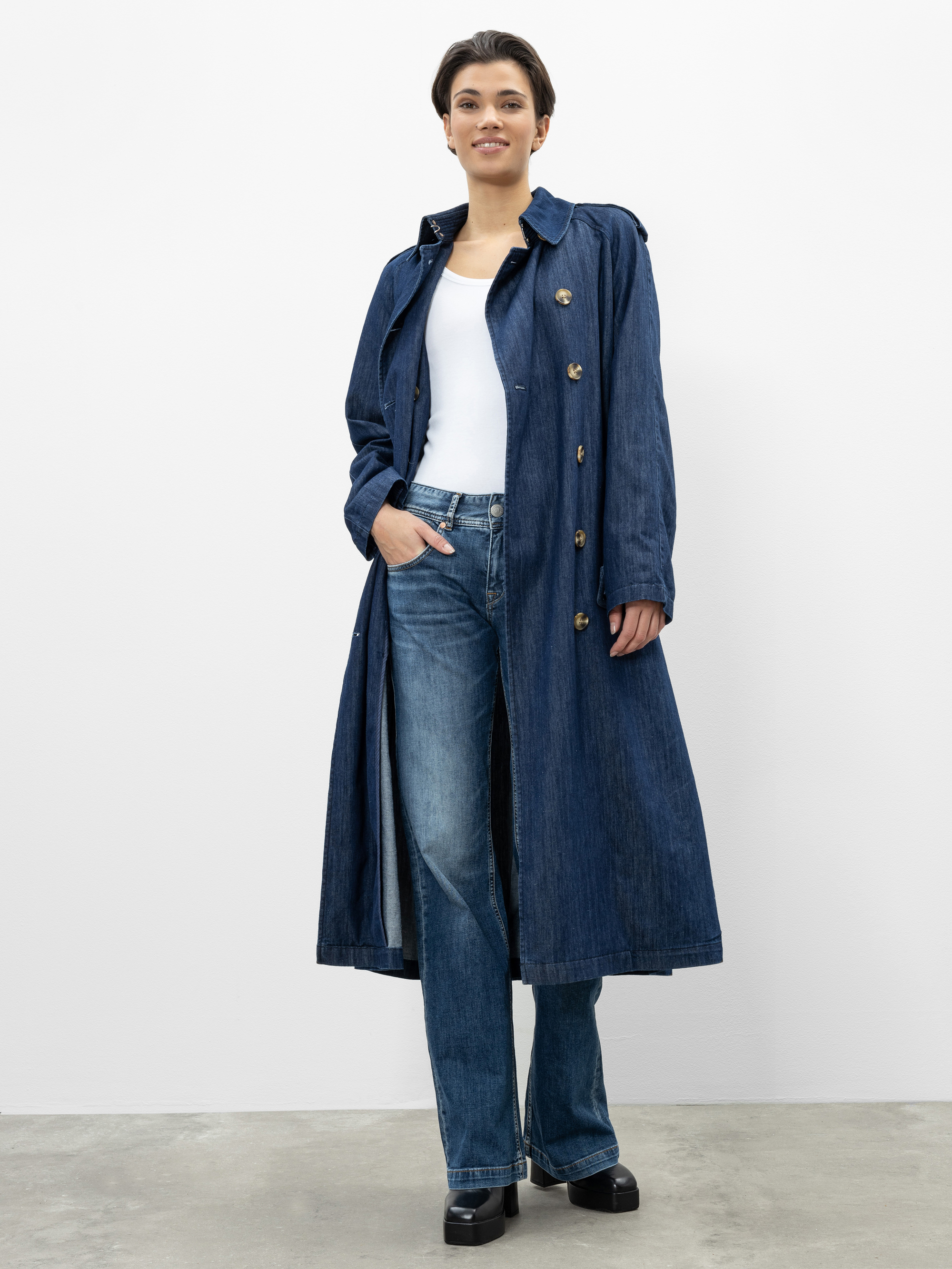 Eine Person, die einen langen blauen Denim-Trenchcoat über einem weißen Oberteil und eine weit geschnittene Jeans mit schwarzen Plateauschuhen trägt, steht lächelnd vor einem schlichten weißen Hintergrund.