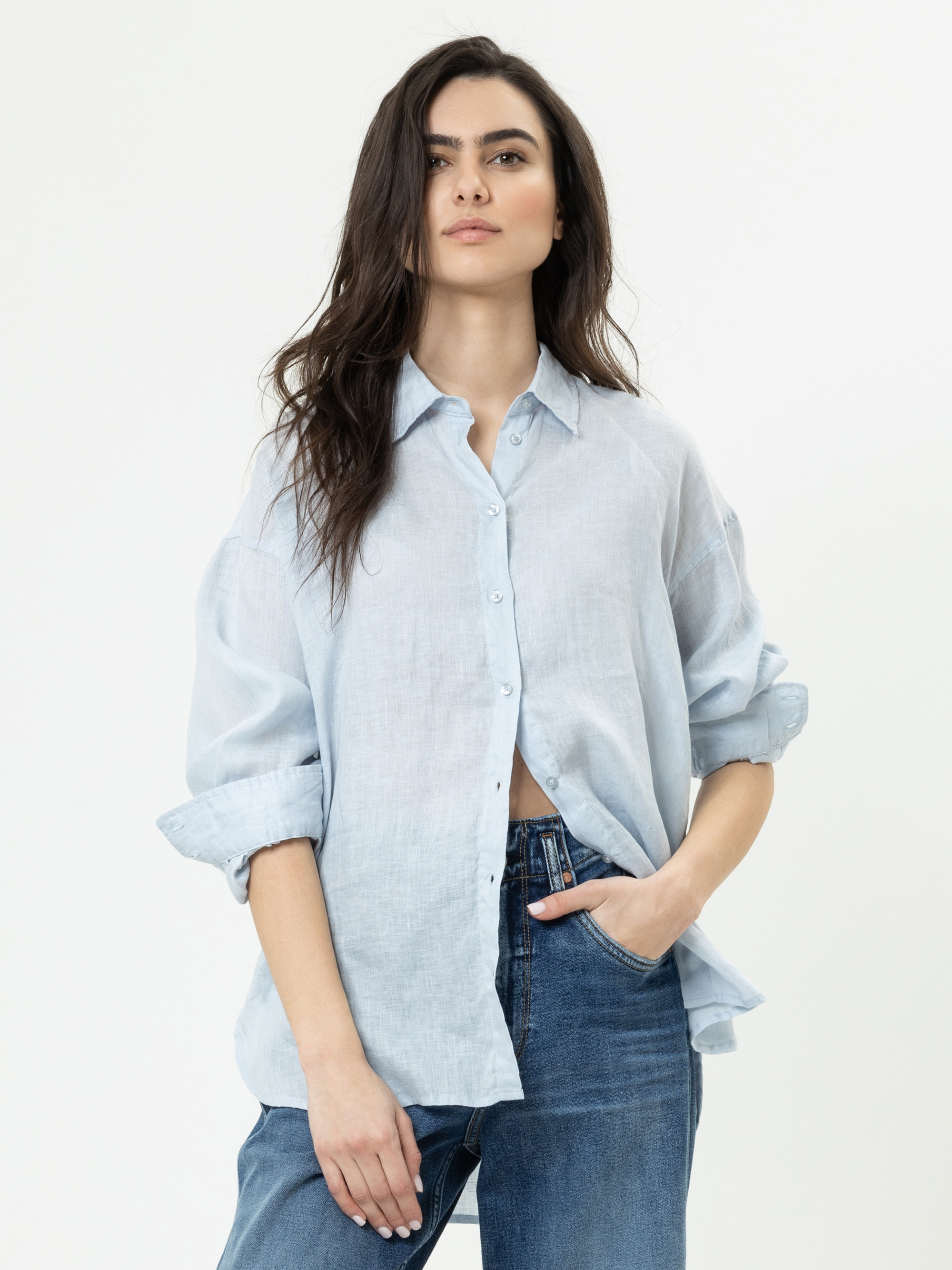 Eine Frau mit langen dunklen Haaren trägt ein hellblaues Button-up-Hemd mit hochgekrempelten Ärmeln und blaue Jeans. Sie steht vor einem schlichten weißen Hintergrund und hat eine Hand in der Tasche.