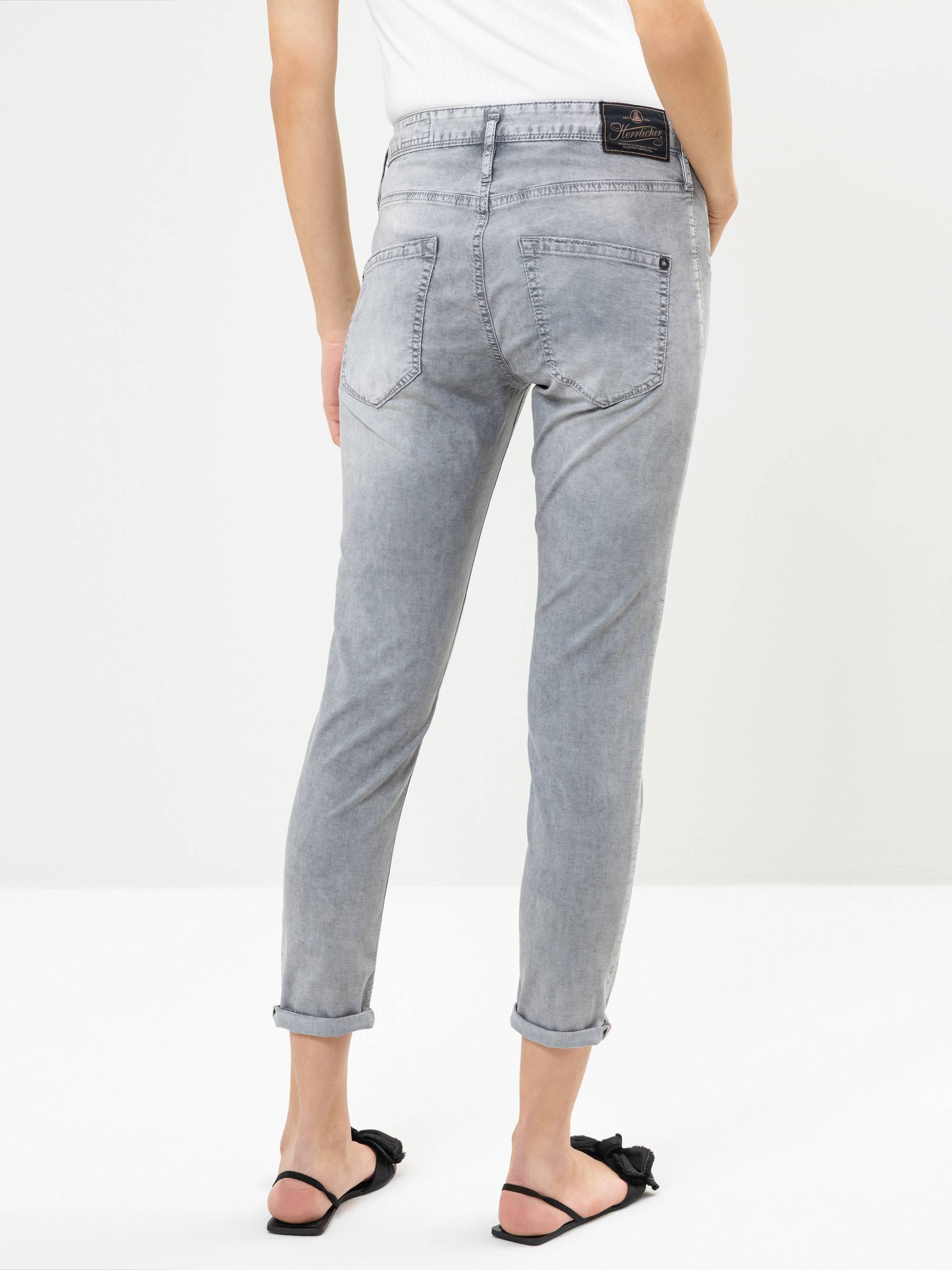 Person in hellgrauer, leicht verblasster Jeans mit hochgekrempelten Manschetten und einem weißen Oberteil, von hinten gesehen. Außerdem trägt sie schwarze Sandalen mit Schleife. Der Hintergrund ist einfarbig weiß.