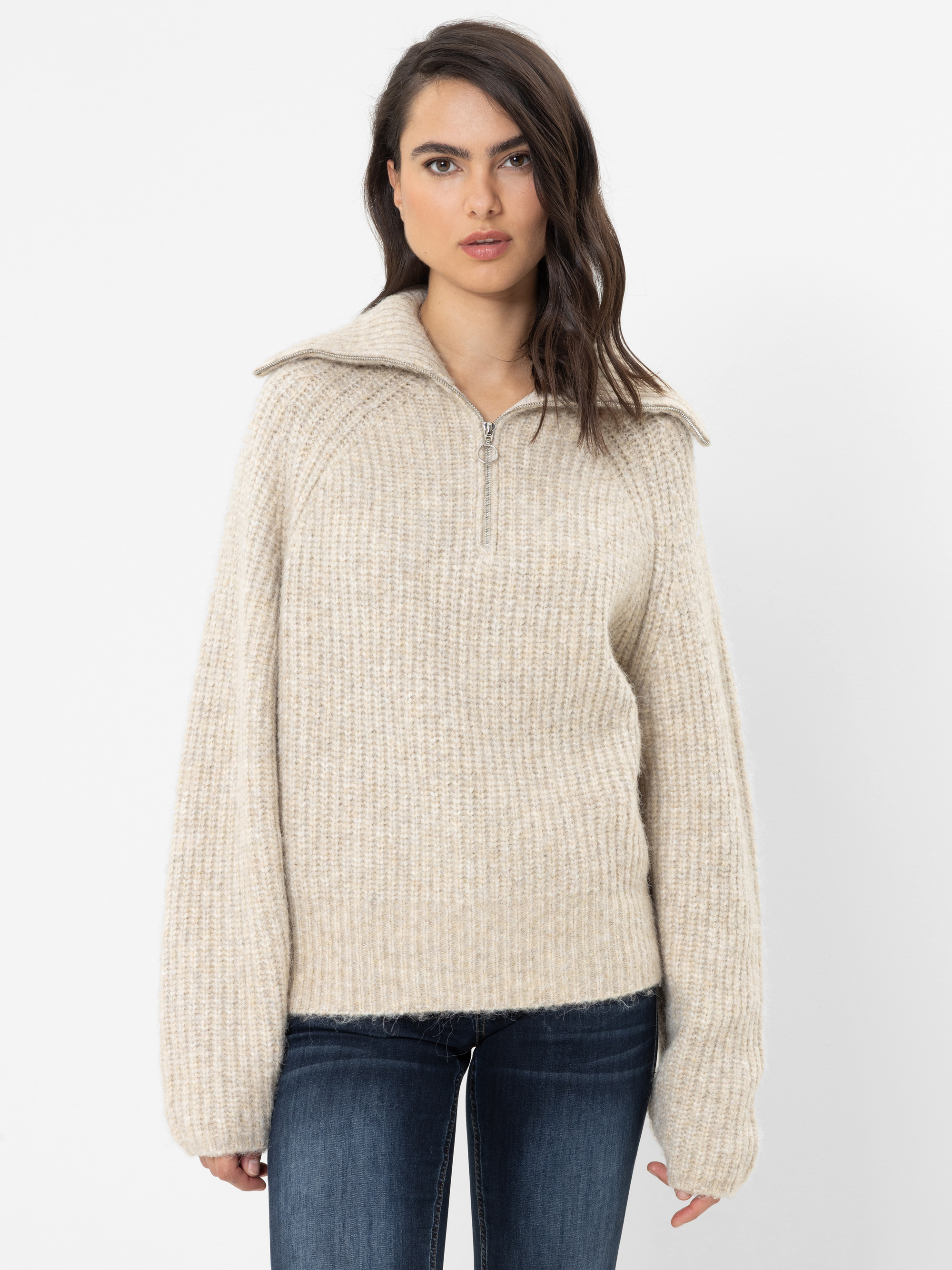 Eine Frau mit langen, dunklen Haaren trägt einen cremefarbenen, gerippten Strickpullover mit halbem Reißverschluss und eine blaue Jeans und steht vor einem schlichten weißen Hintergrund.