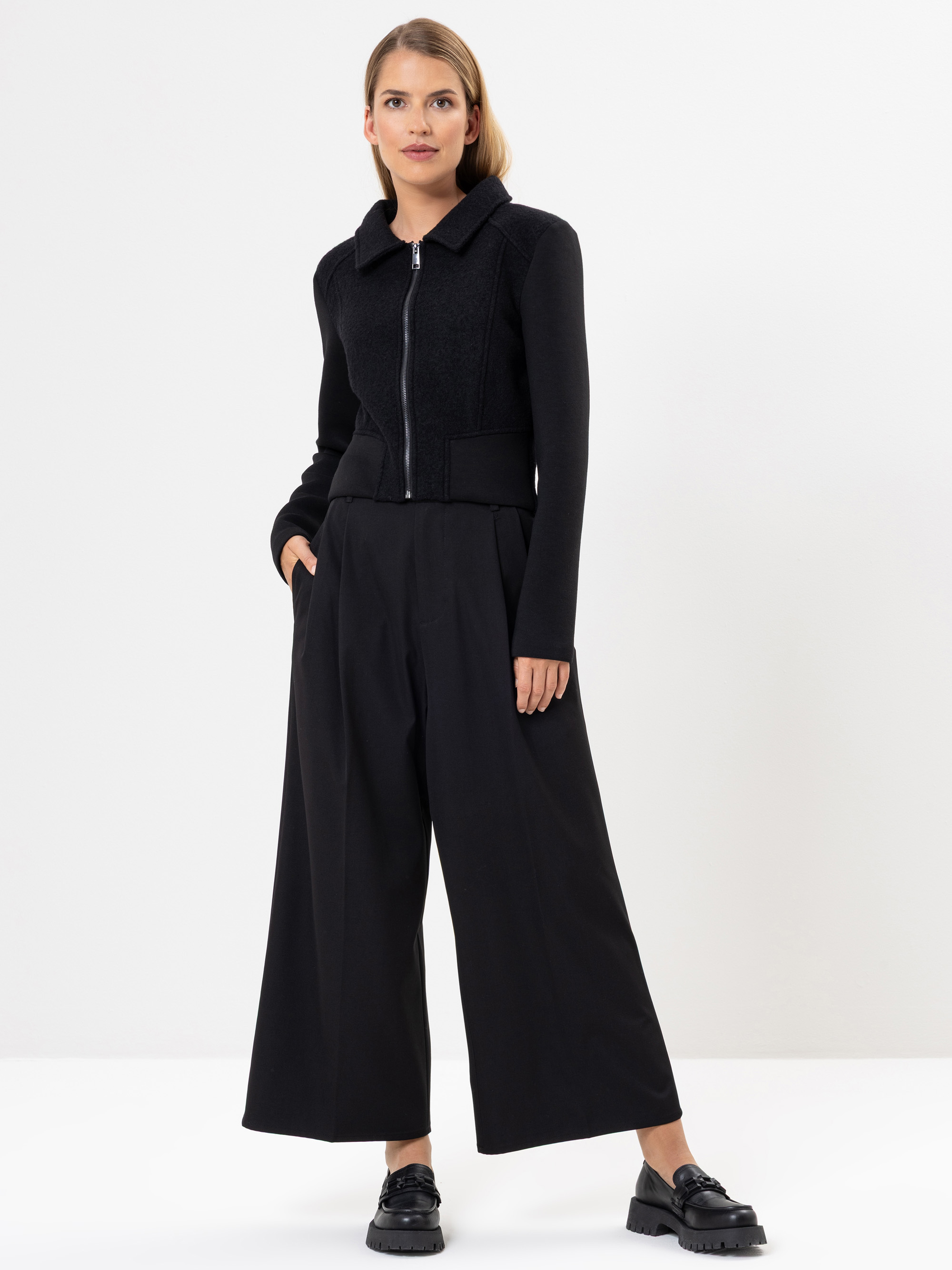 Eine Frau steht vor einem schlichten weißen Hintergrund und trägt eine schwarze Jacke mit Reißverschluss, eine weit geschnittene schwarze Hose und schwarze Schuhe. Eine Hand steckt in ihrer Tasche, und sie blickt selbstbewusst in die Kamera.
