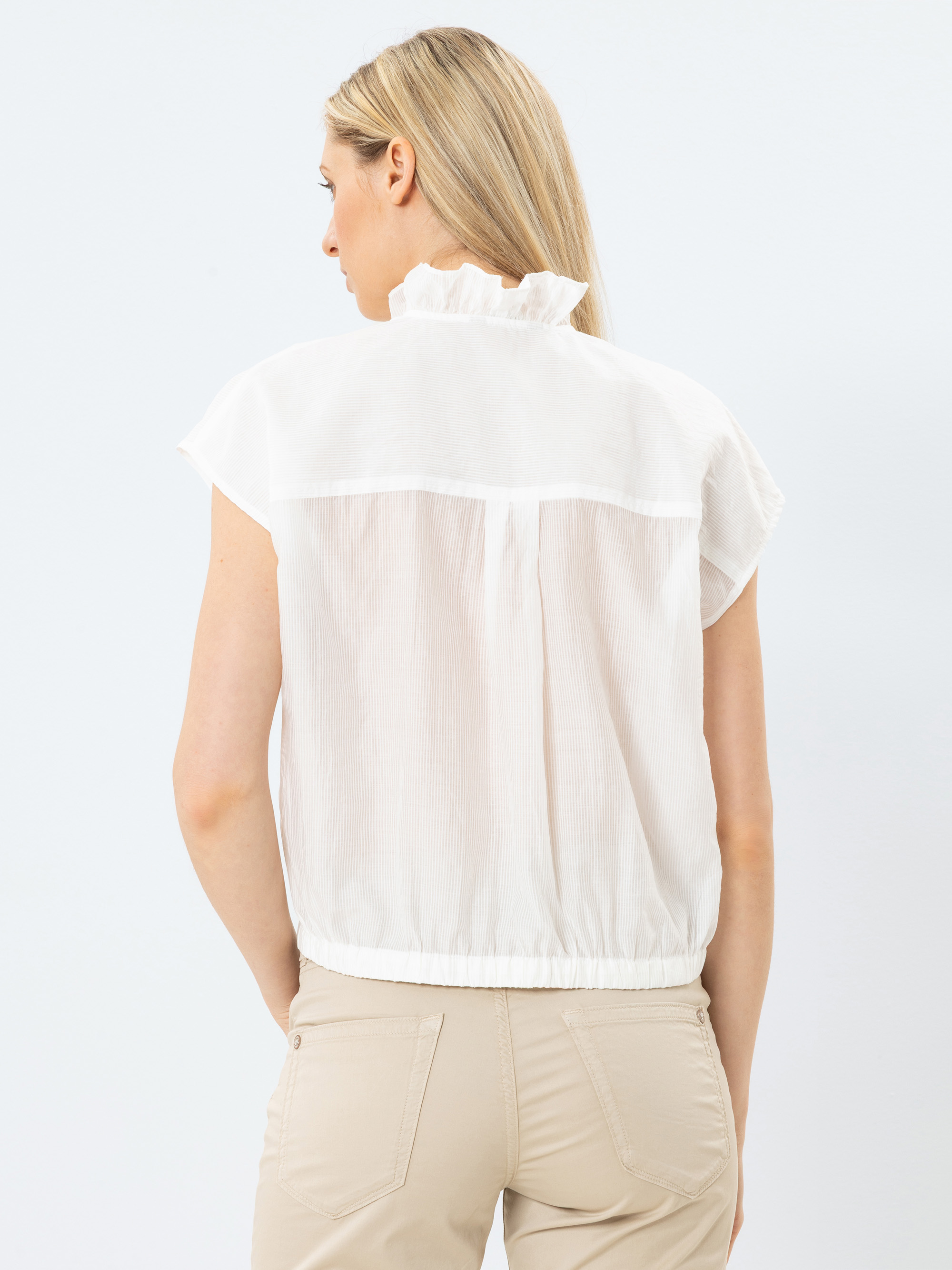 Eine Frau mit langen blonden Haaren, von hinten gesehen, trägt eine kurzärmelige, weiße Bluse mit Rüschenkragen und eine hellbeige Hose vor einem schlichten weißen Hintergrund.