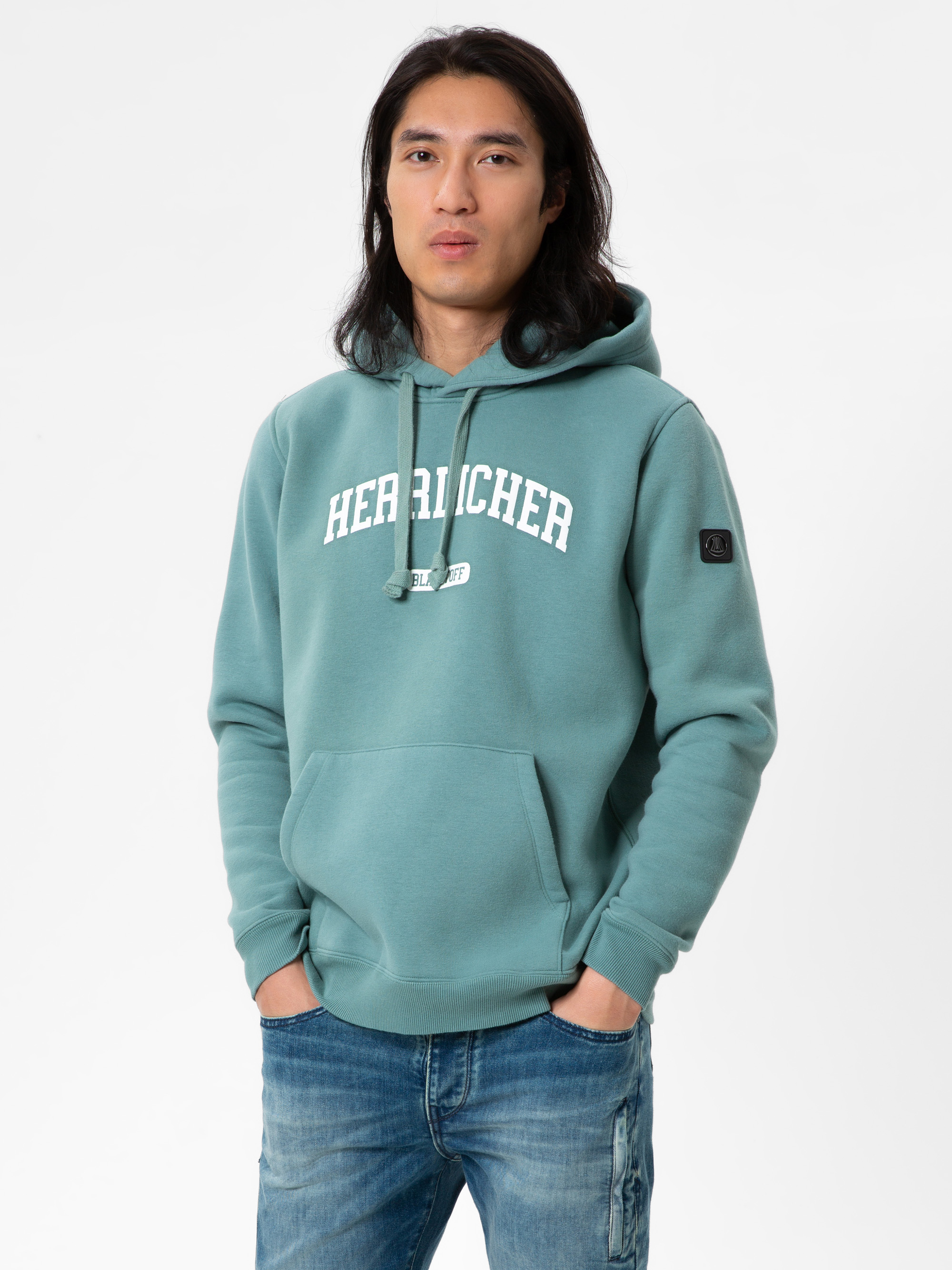 Eine Person mit langen dunklen Haaren, die ein blaugrünes Kapuzensweatshirt mit dem Aufdruck HERZLICHER 03 auf der Vorderseite und blaue Jeans trägt, steht vor einem schlichten weißen Hintergrund.