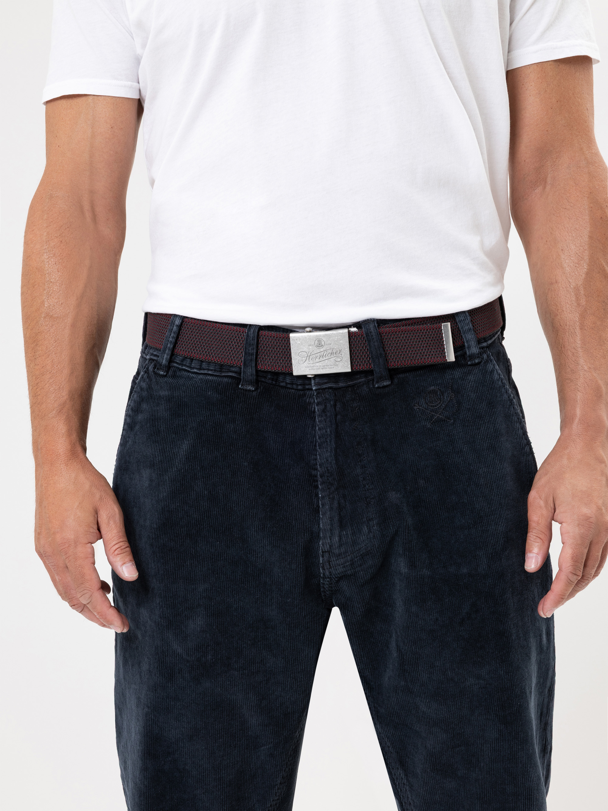 Eine Person, die ein weißes T-Shirt, eine dunkelblaue Kordhose und einen dunklen Gürtel mit einer silbernen Schnalle trägt. Nur der Oberkörper und die Oberschenkel sind vor einem einfarbigen Hintergrund zu sehen.