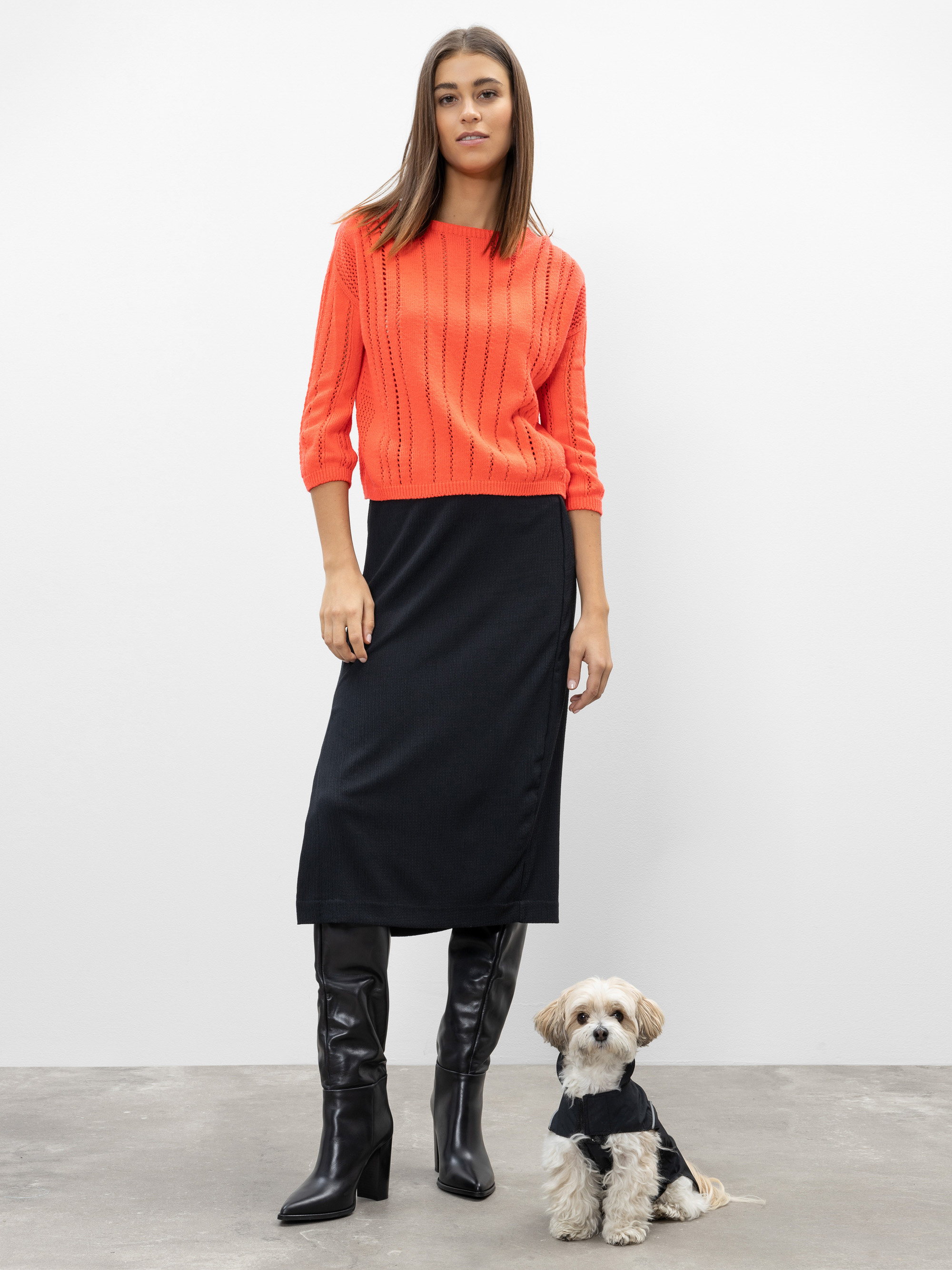 Eine Frau in einem leuchtend orangefarbenen Pullover, einem schwarzen Midirock und schwarzen Stiefeln steht vor einem schlichten Hintergrund. Ein kleiner Hund in einem schwarzen Pullover sitzt zu ihren Füßen und schaut nach vorne.