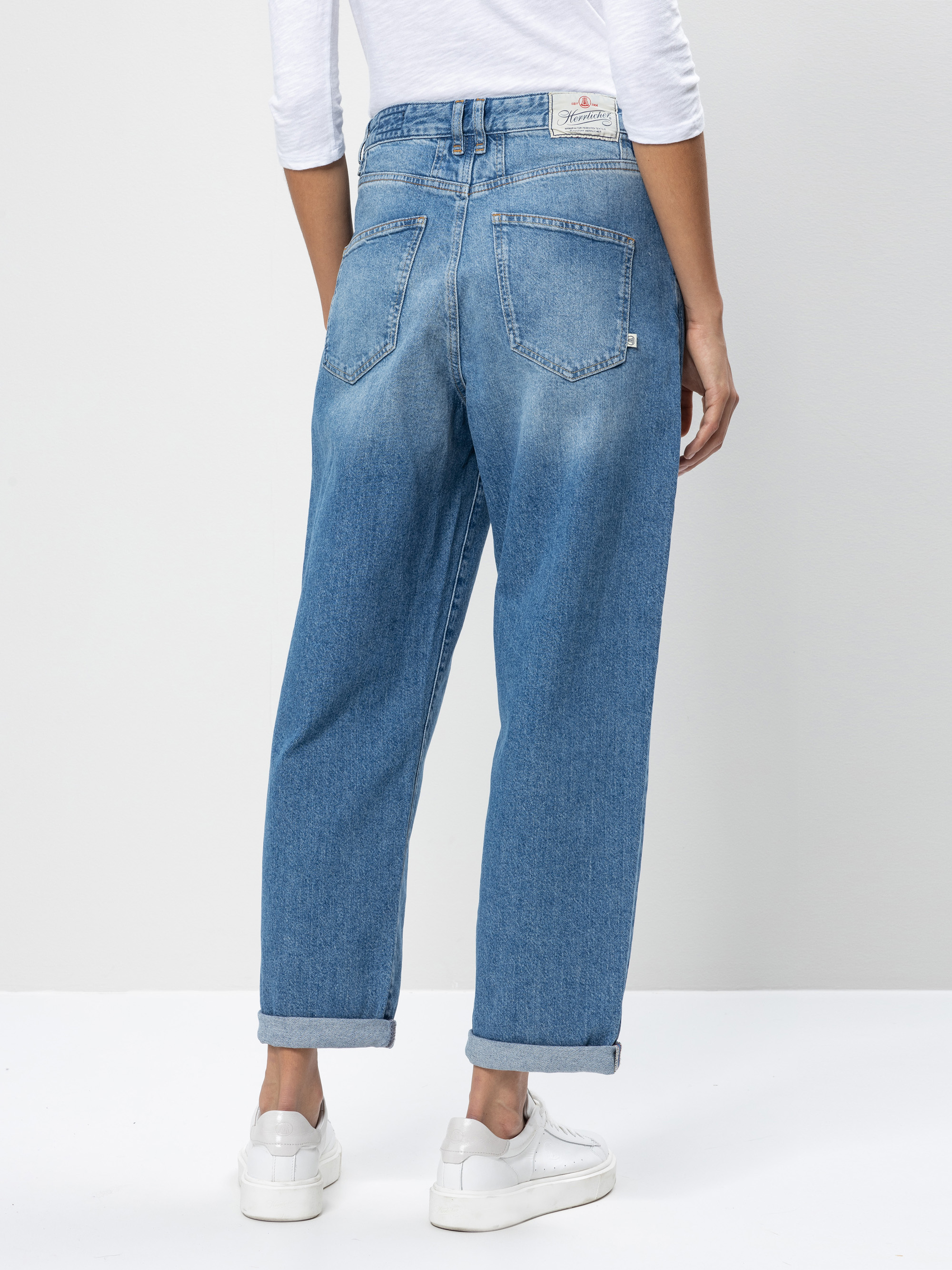 Eine Person, die eine hellblaue Jeans mit hoher Taille, ein weißes langärmeliges Oberteil und weiße Turnschuhe trägt, steht mit dem Gesicht nach unten vor einem einfarbigen hellgrauen Hintergrund.