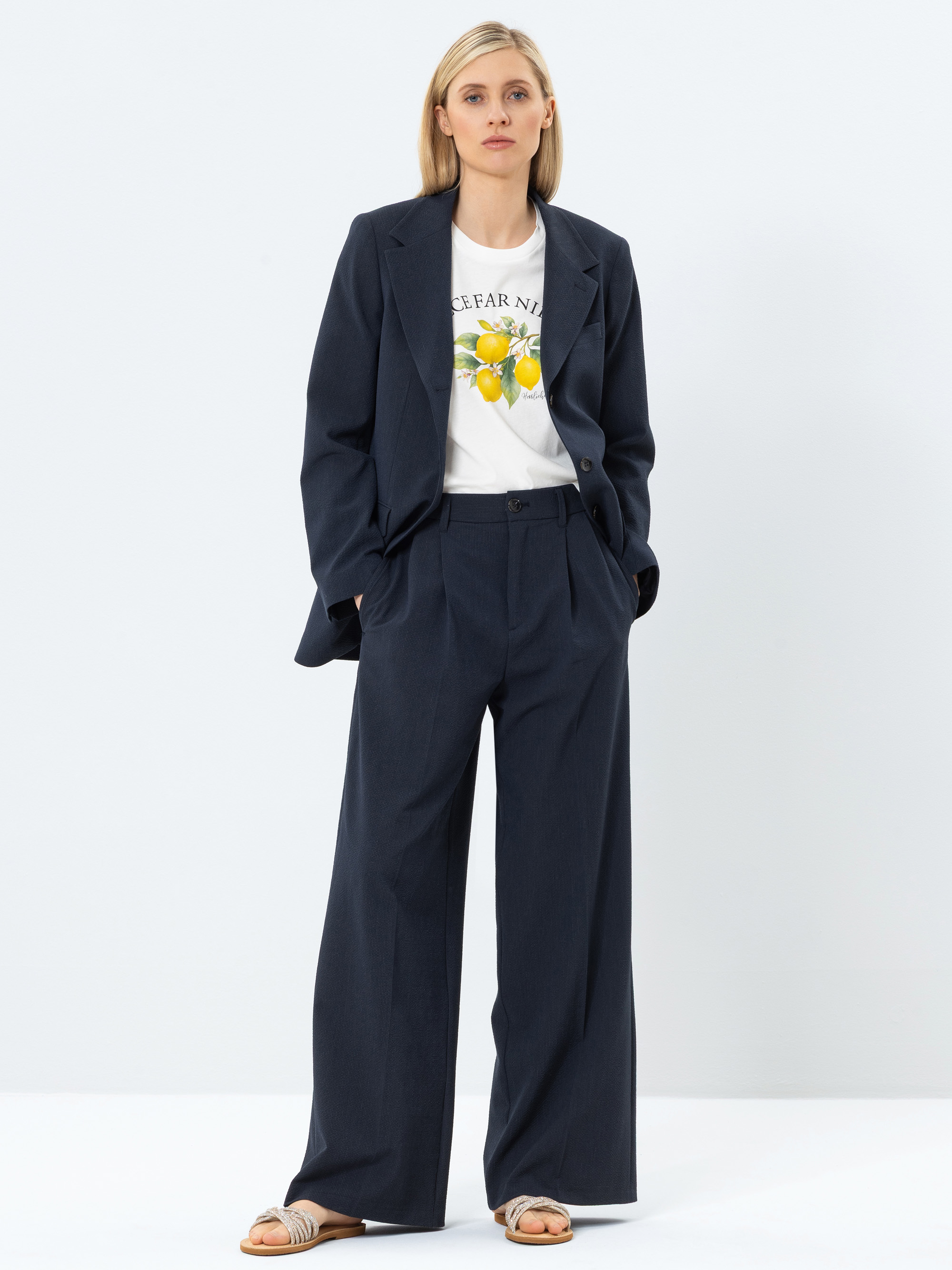 Eine Frau steht vor einem schlichten Hintergrund und trägt einen dunkelblauen Anzug in Übergröße, ein weißes T-Shirt mit Zitronenmotiven und beigefarbene Sandalen. Sie hat blondes Haar, die Hände in den Hosentaschen und einen neutralen Gesichtsausdr