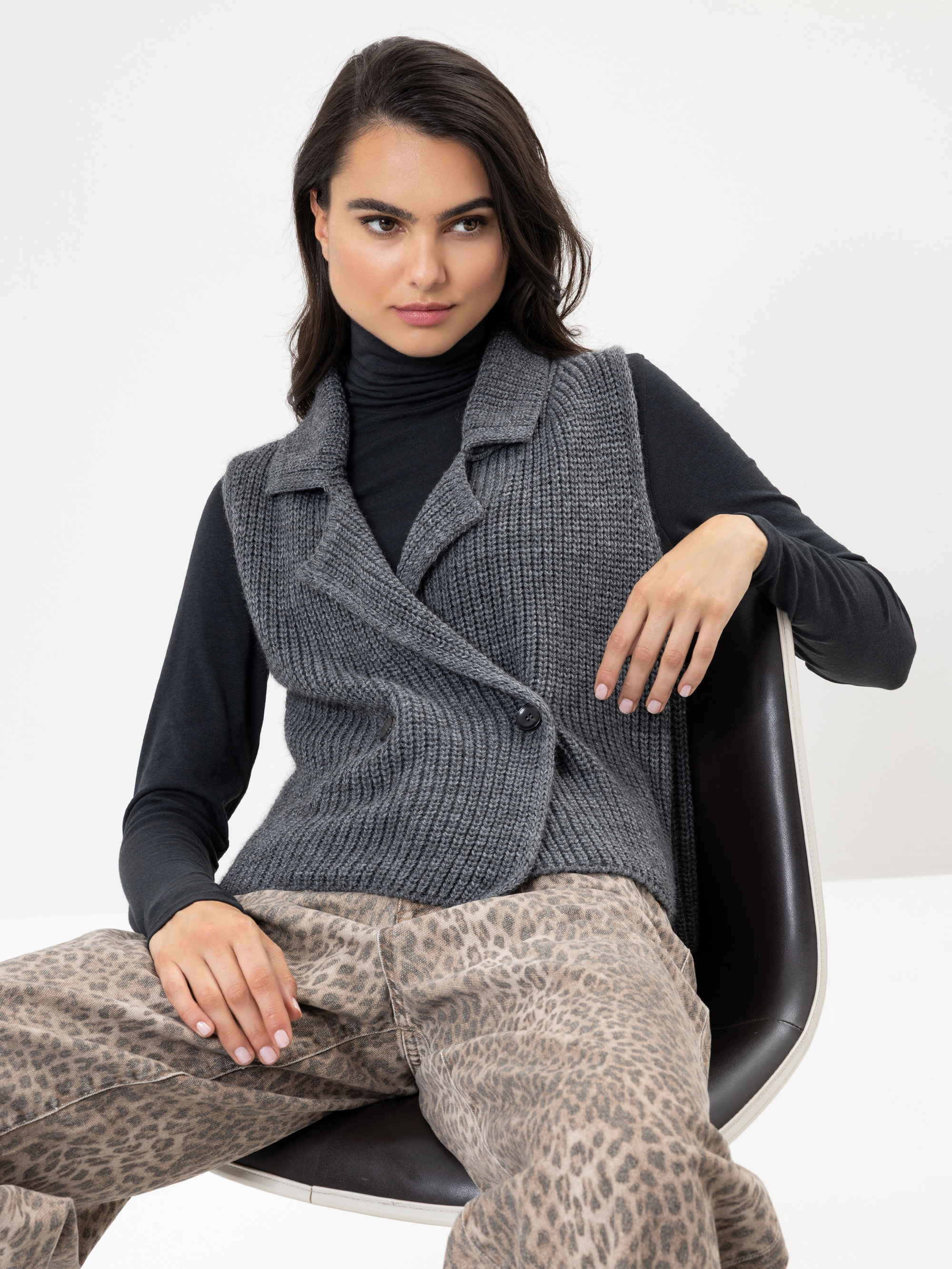 Eine Frau sitzt auf einem modernen schwarzen Stuhl, trägt eine graue Strickweste über einem schwarzen Rollkragenpullover und eine beigefarbene Hose mit Leopardenmuster. Sie hat langes dunkles Haar und einen neutralen Gesichtsausdruck, eine Hand stützt