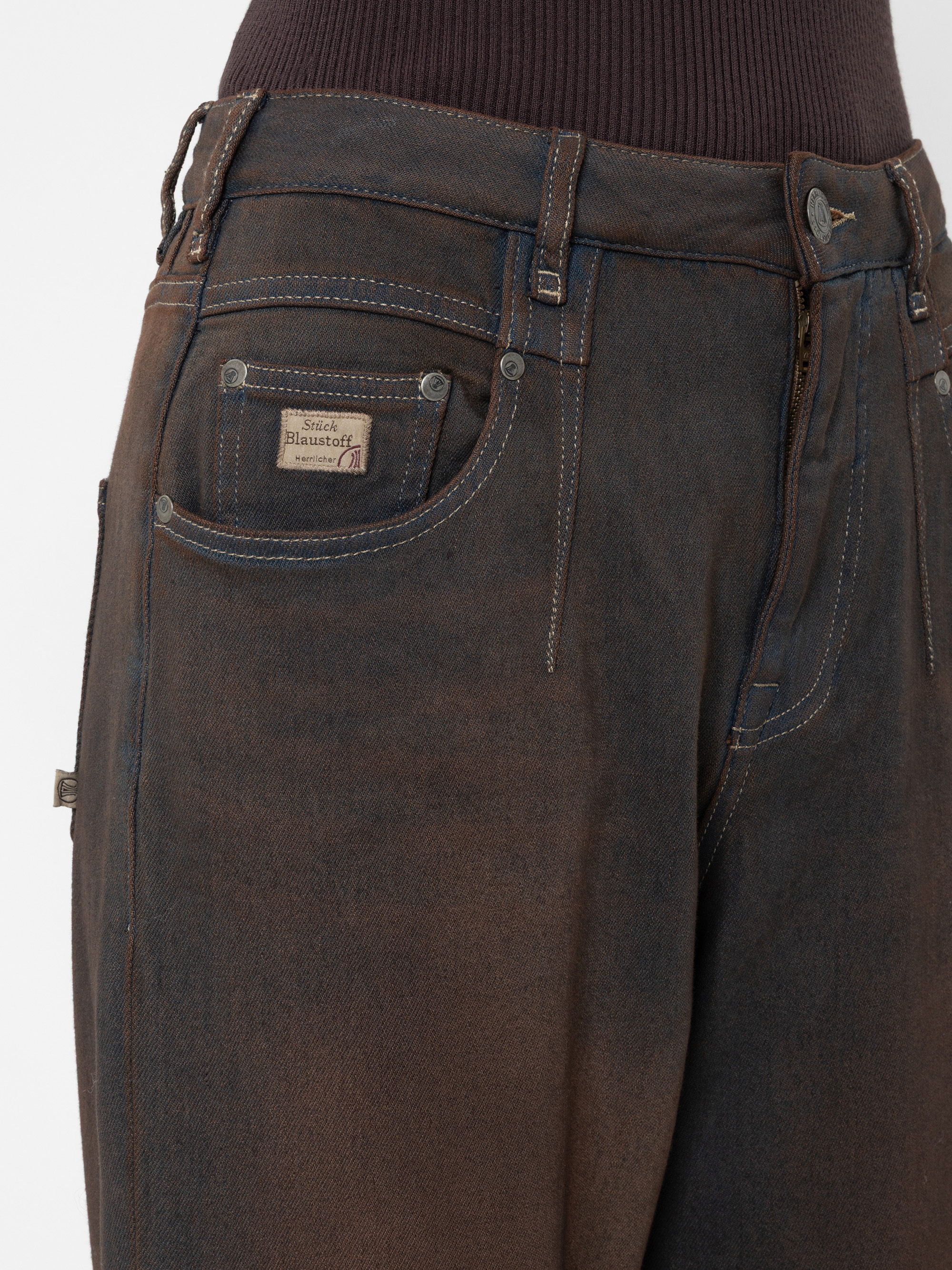 Nahaufnahme einer braunen Jeans mit sichtbaren Nähten, einer Vordertasche, einer Münztasche mit Etikett und Metallnieten. Die Jeans wird mit einem gerippten, dunklen Oberteil kombiniert.