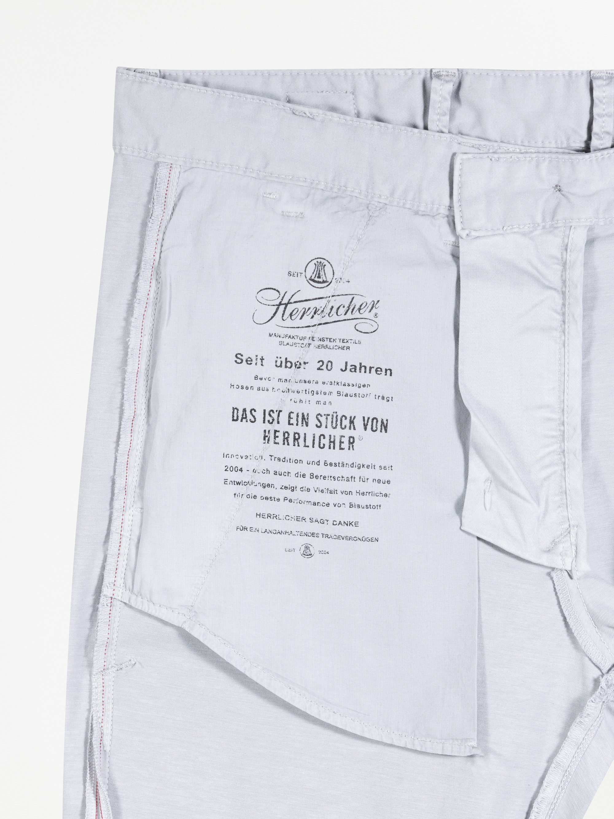 Hellgraue Hose mit umgedrehter Innenseite. Auf der Innentasche ist ein Etikett mit dem Markennamen Herrlicher und einem deutschen Text über die Qualität und Geschichte der Marke aufgedruckt. Einige Nähte und Taschendetails sind sichtbar.