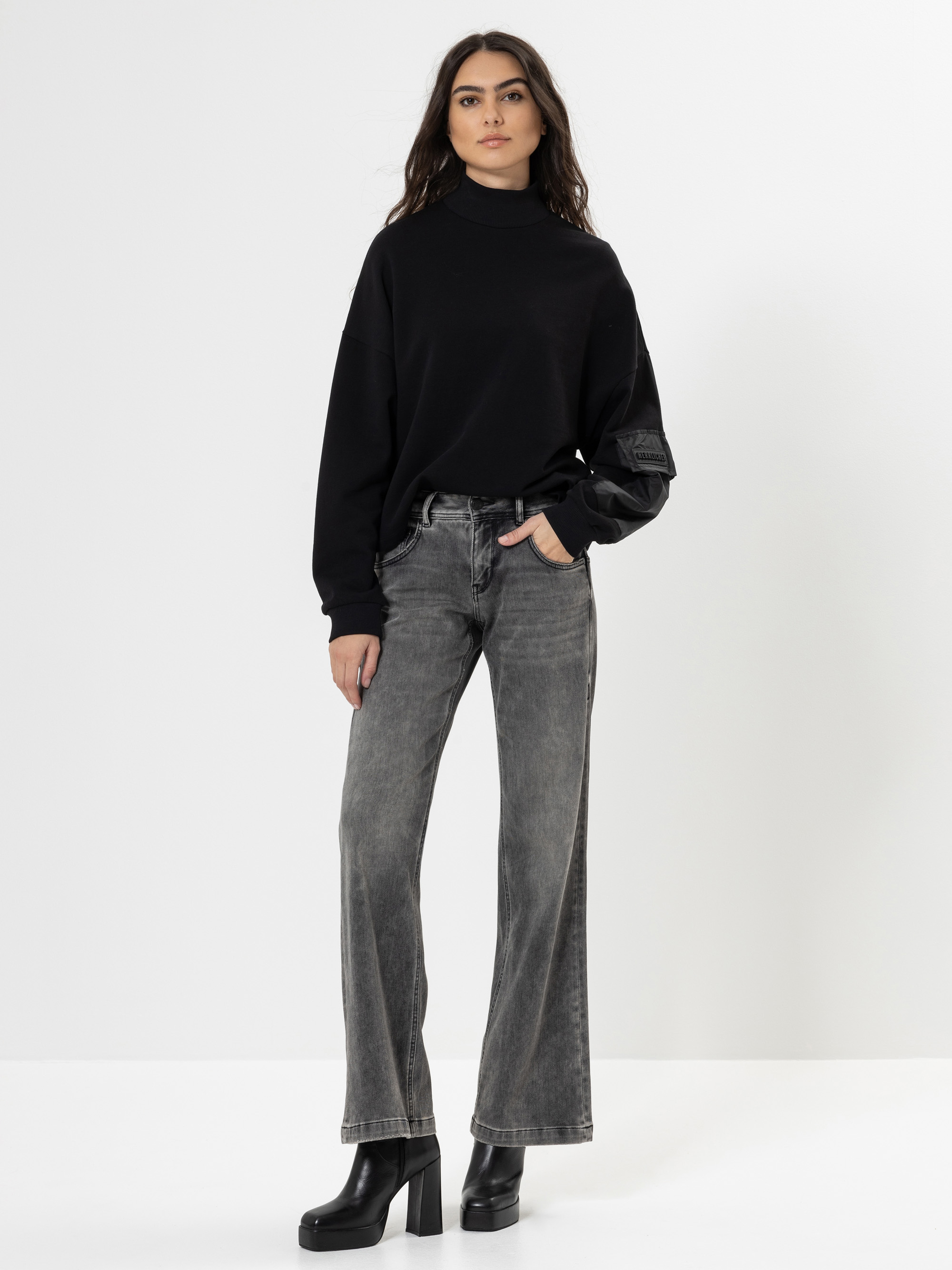 Eine Frau steht vor einem schlichten weißen Hintergrund und trägt einen schwarzen Rollkragenpullover, verblichene graue Jeans mit weitem Bein und schwarze Plateauschuhe. Sie hat langes dunkles Haar und eine Hand in der Tasche.