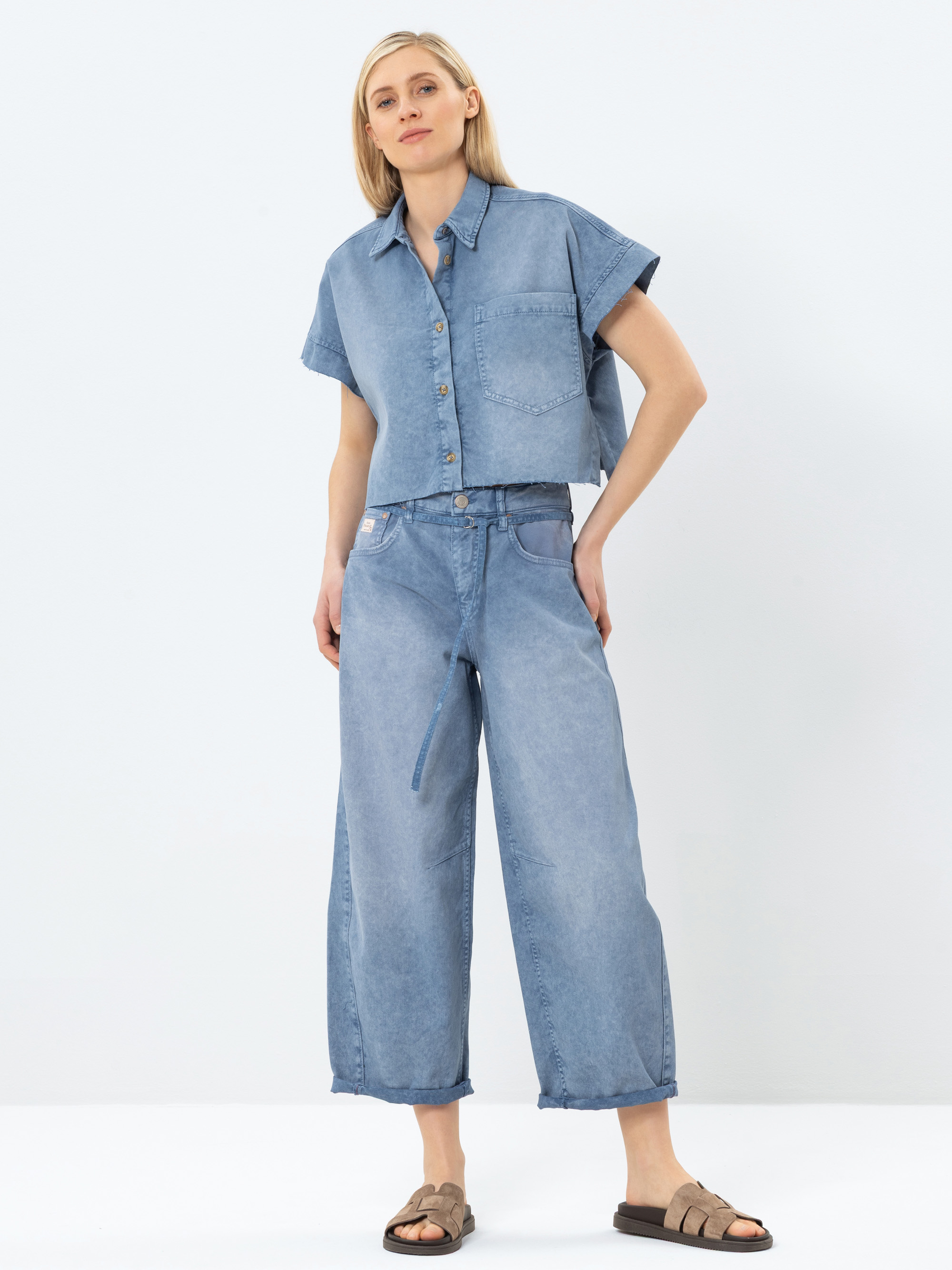 Eine Person steht vor einem schlichten Hintergrund und trägt ein kurzärmeliges, hochgeknöpftes Jeanshemd und Jeans mit weitem Bein. Sie hat helles Haar und trägt braune Sandalen. Ihre Hände stecken in den Taschen und sie sieht entspannt aus.