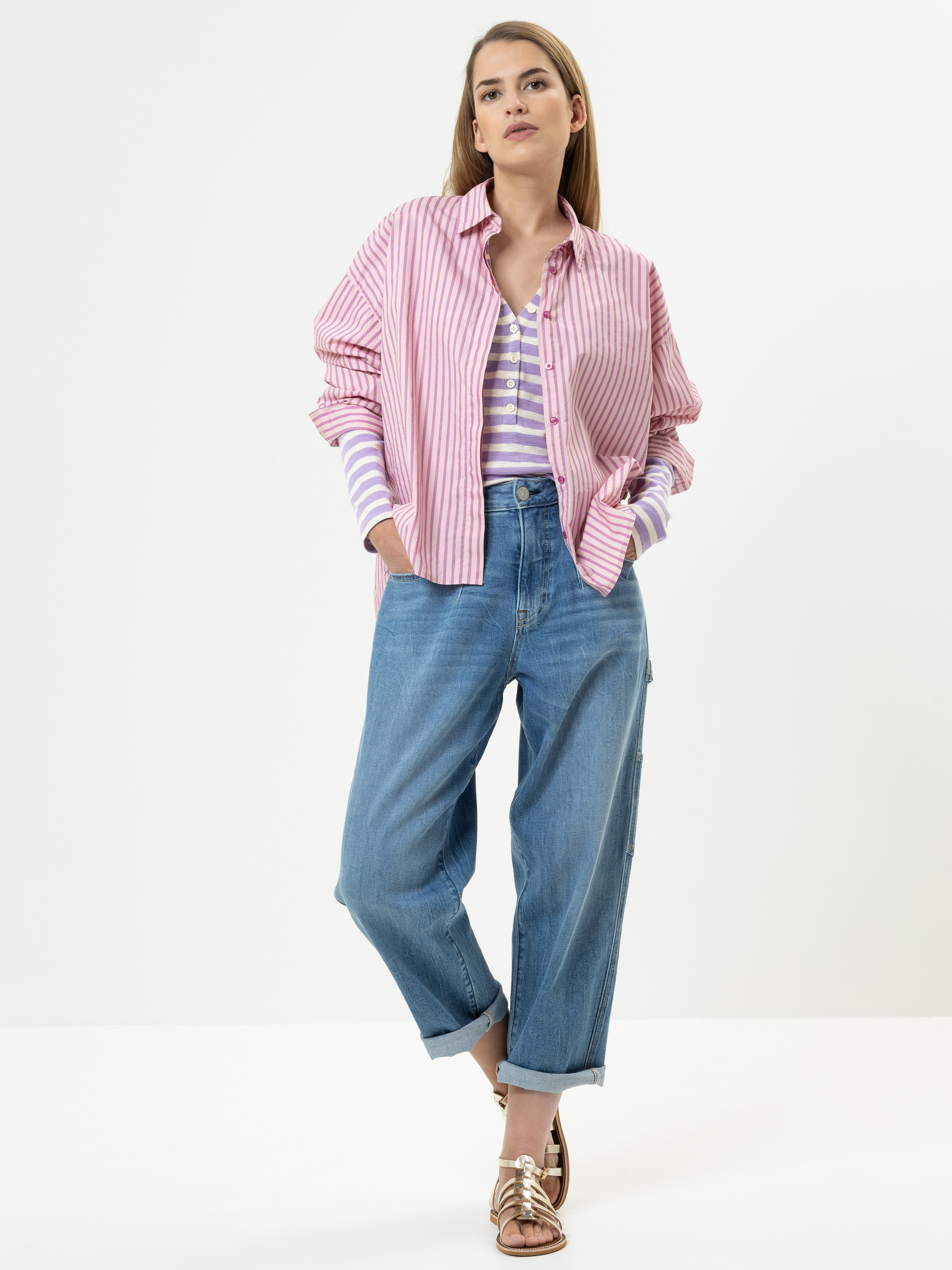 Eine Frau steht vor einem weißen Hintergrund und trägt ein rosa gestreiftes Hemd über einem lavendelfarbenen gestreiften Oberteil, weite Bluejeans mit aufgerollten Manschetten und Metallic-Sandalen, die Hände in den Taschen und einen neutralen Blick.