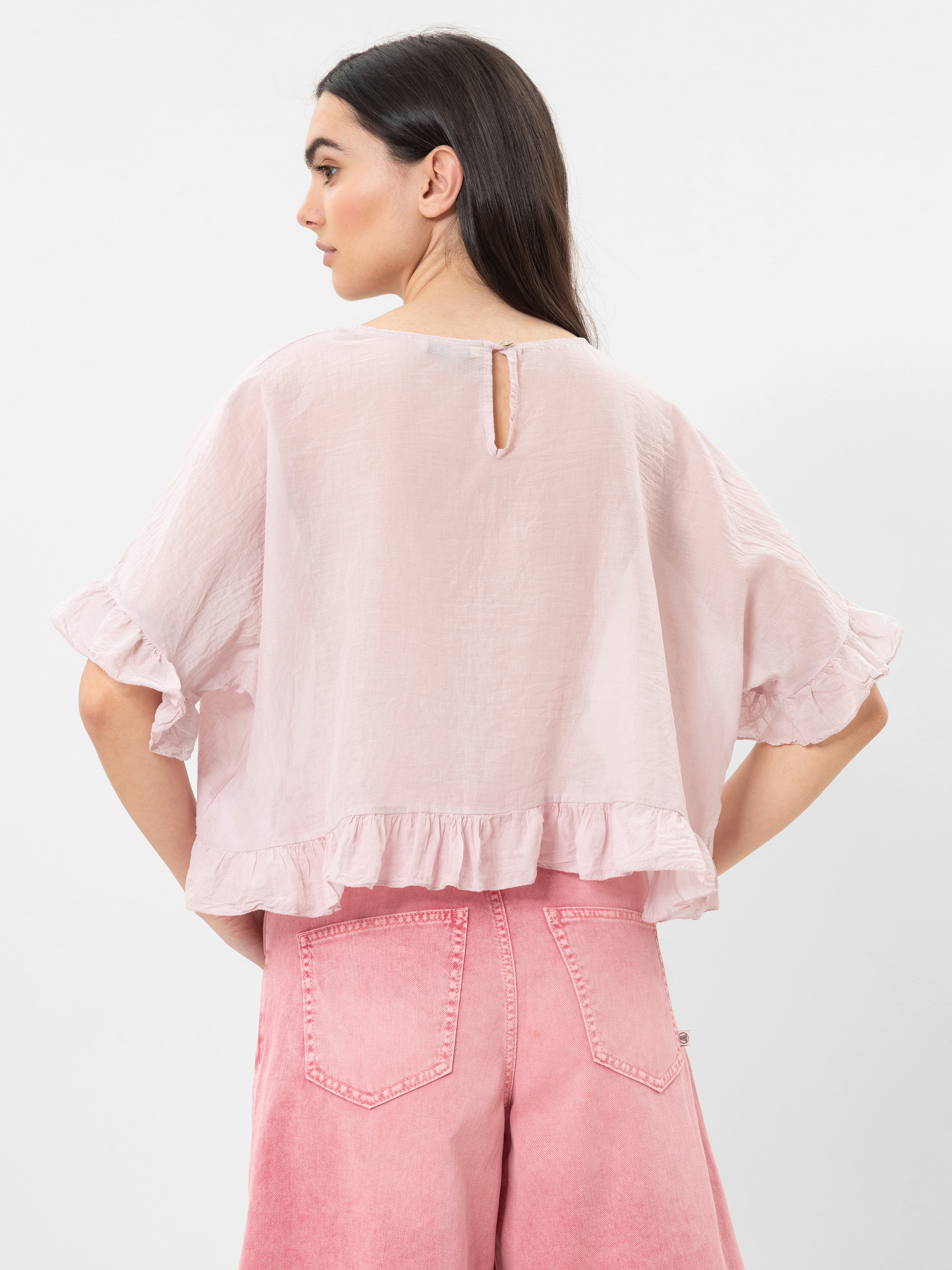 Eine Frau mit langen dunklen Haaren trägt eine lockere, hellrosa Bluse mit gerüschten Ärmeln und Saum, gepaart mit einer hochgeschnittenen rosa Jeans. Sie ist abgewandt und zeigt die Rückseite ihres Outfits vor einem schlichten Hintergrund.