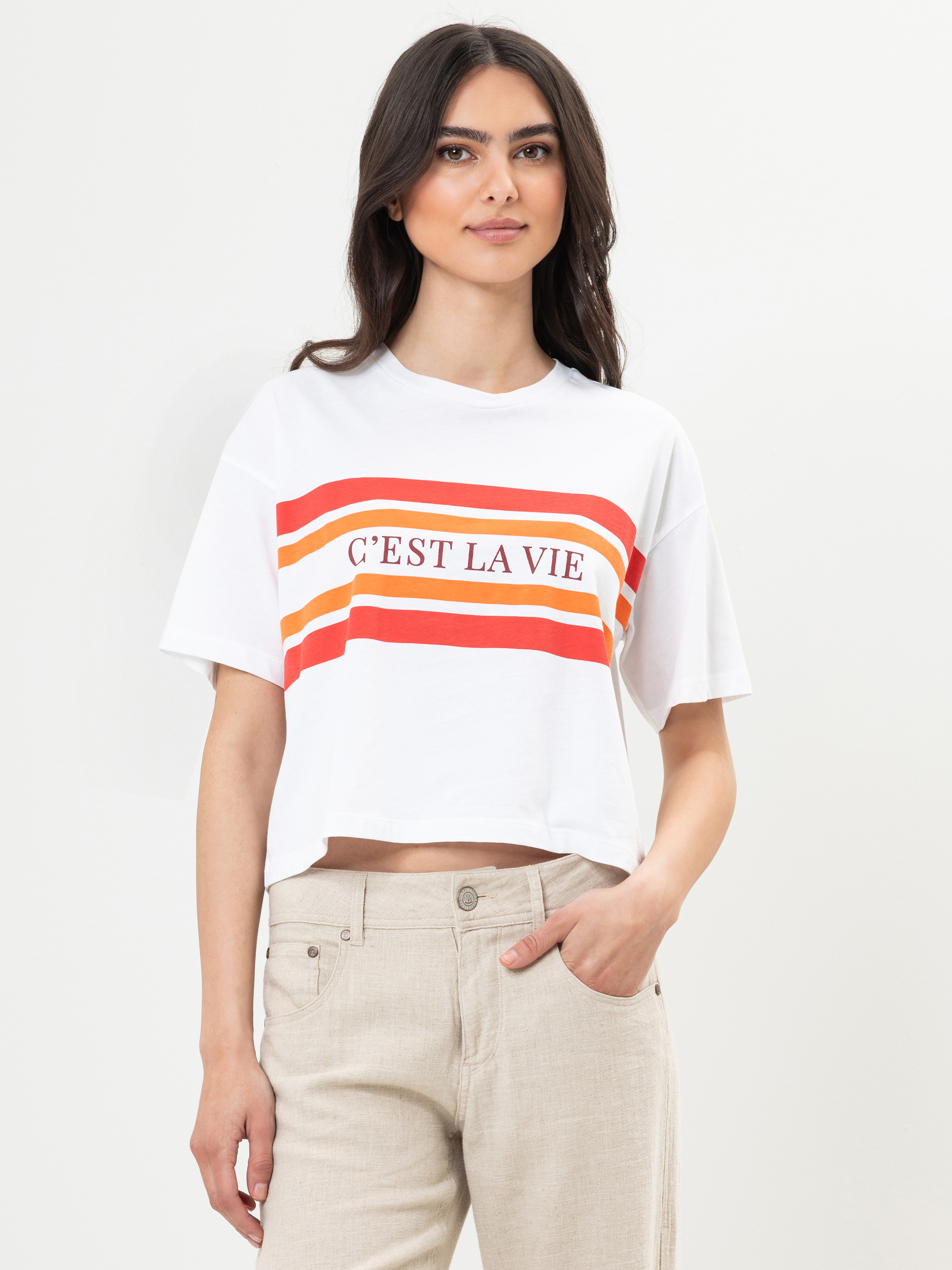 Eine Frau mit langen dunklen Haaren trägt ein weißes T-Shirt mit roten und orangefarbenen Streifen und dem Schriftzug CEST LA VIE auf der Vorderseite, gepaart mit einer beigen Hose, vor einem schlichten weißen Hintergrund.