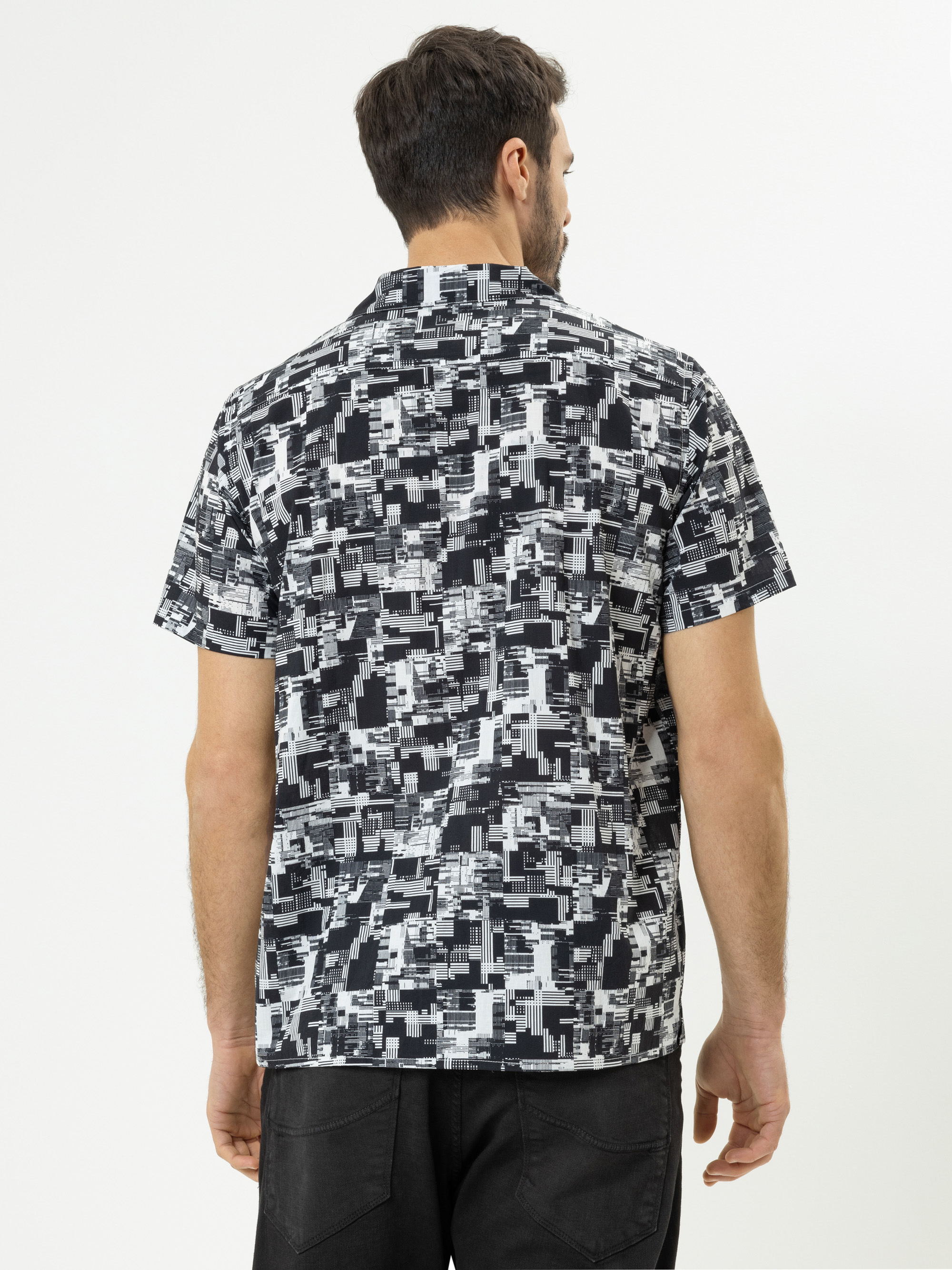 Ein Mann steht mit dem Rücken zur Kamera und trägt ein kurzärmeliges Hemd mit einem abstrakten geometrischen Muster in Schwarz, Weiß und Grau sowie eine schwarze Hose vor einem schlichten weißen Hintergrund.