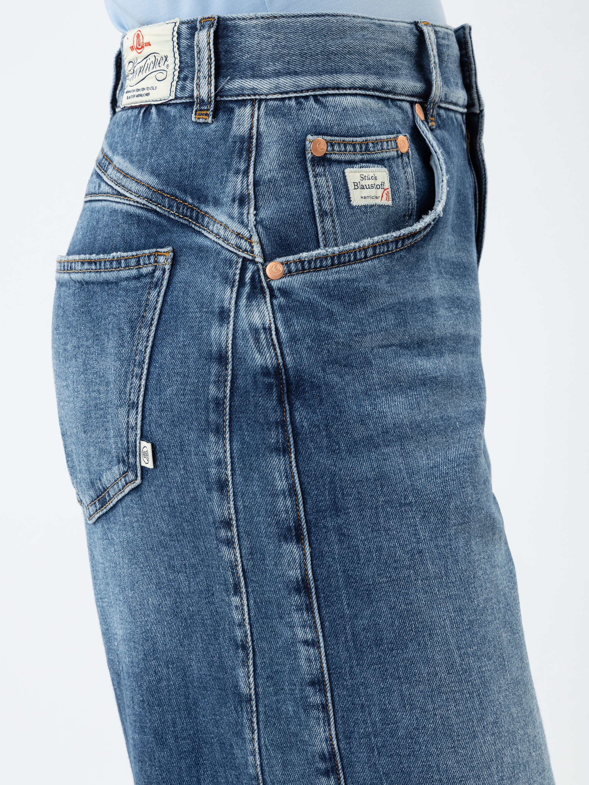 Nahaufnahme einer Person, die eine blaue Denim-Jeans mit sichtbaren Nähten, Nieten und Markenetiketten an Bund und Taschen trägt. Die Jeans hat ein klassisches Five-Pocket-Design.