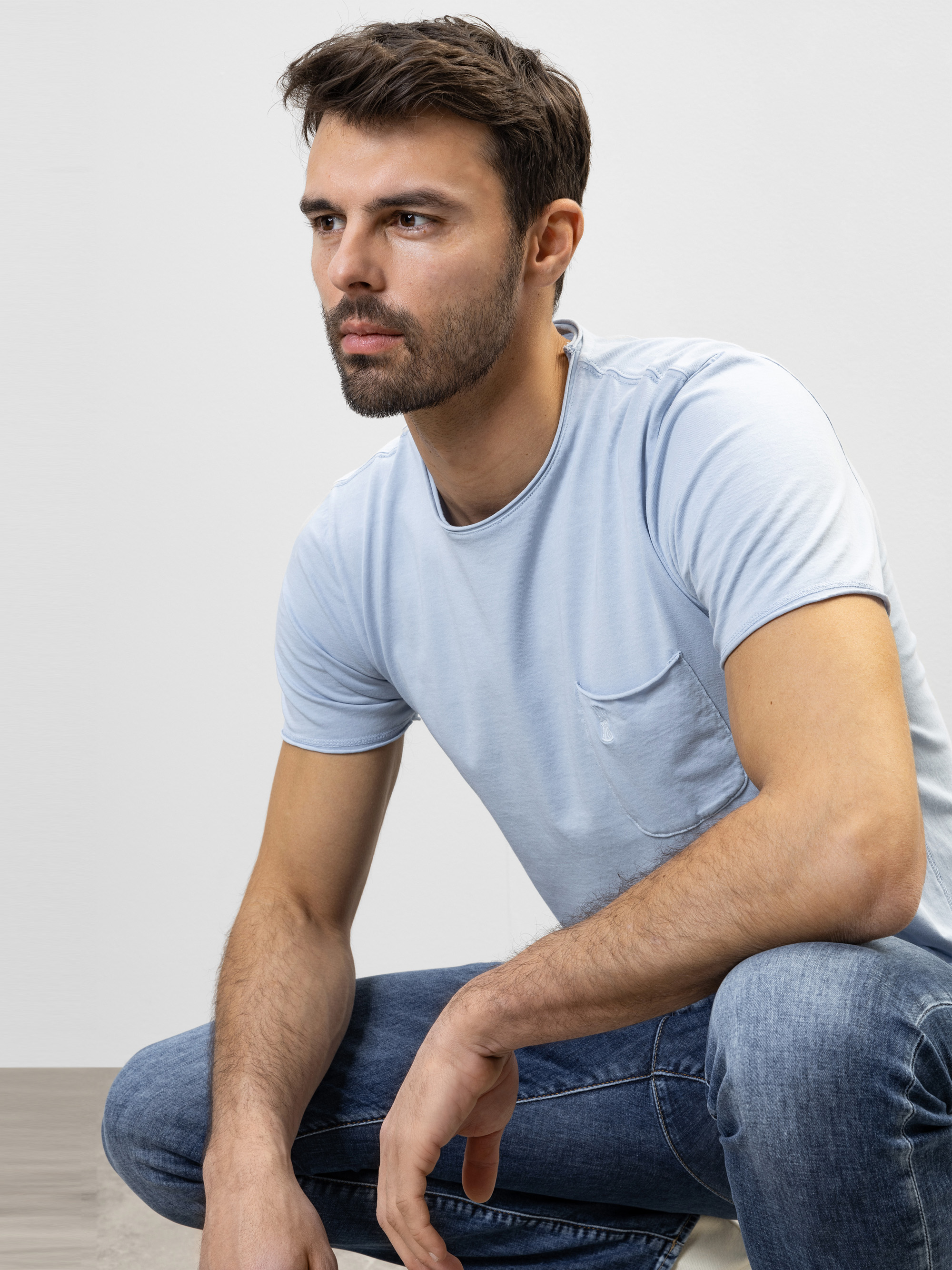 Ein Mann mit kurzen braunen Haaren und Bart, bekleidet mit einem hellblauen T-Shirt und Jeans, sitzt leicht nach vorne gebeugt und schaut nachdenklich vor einem schlichten hellen Hintergrund.
