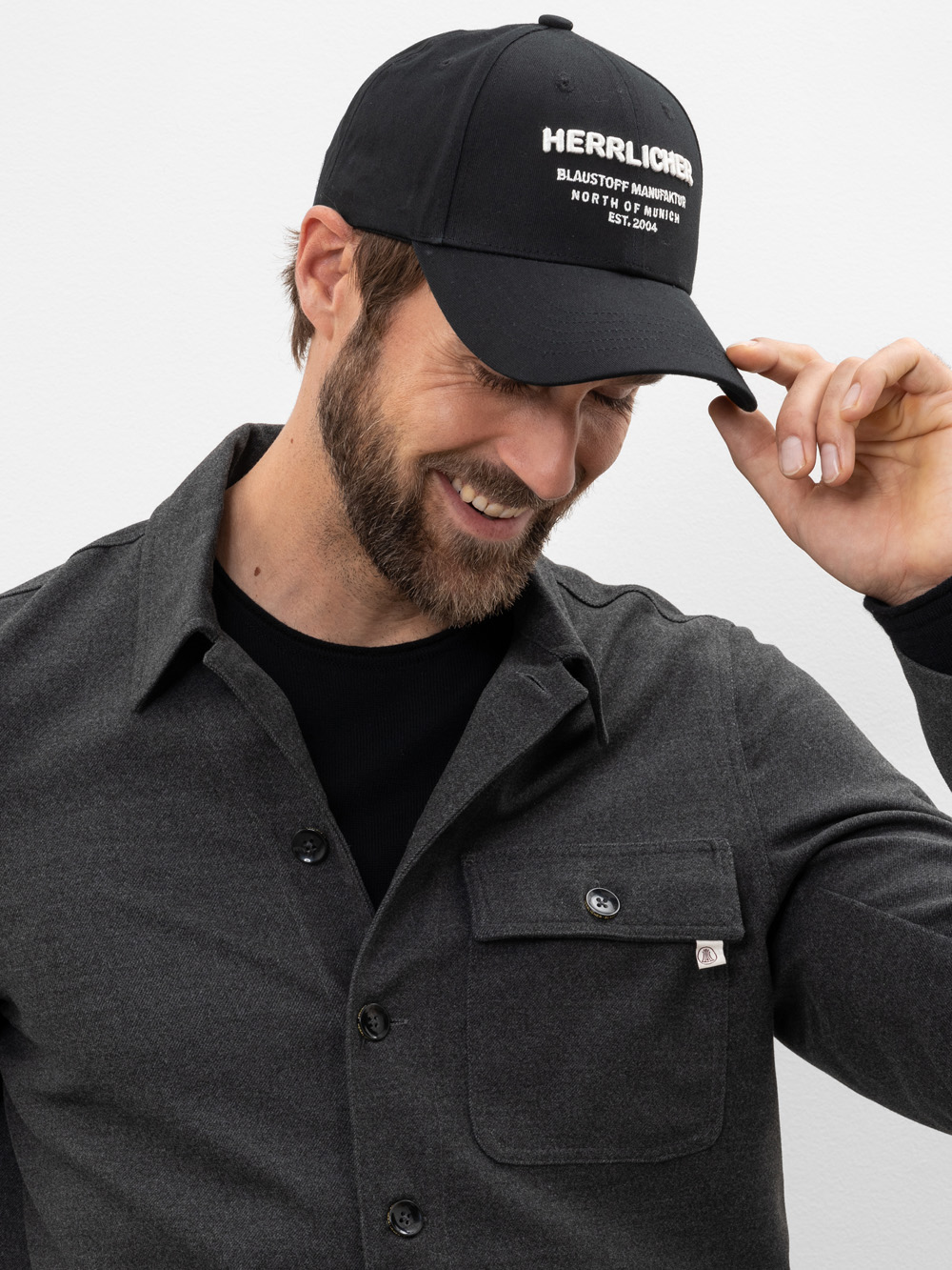 Ein Mann mit Bart lächelt und rückt eine schwarze Mütze zurecht, auf der Herrlicher und andere weiße Schrift zu lesen ist. Er trägt ein dunkles Button-up-Hemd über einem schwarzen T-Shirt vor einem schlichten hellen Hintergrund.