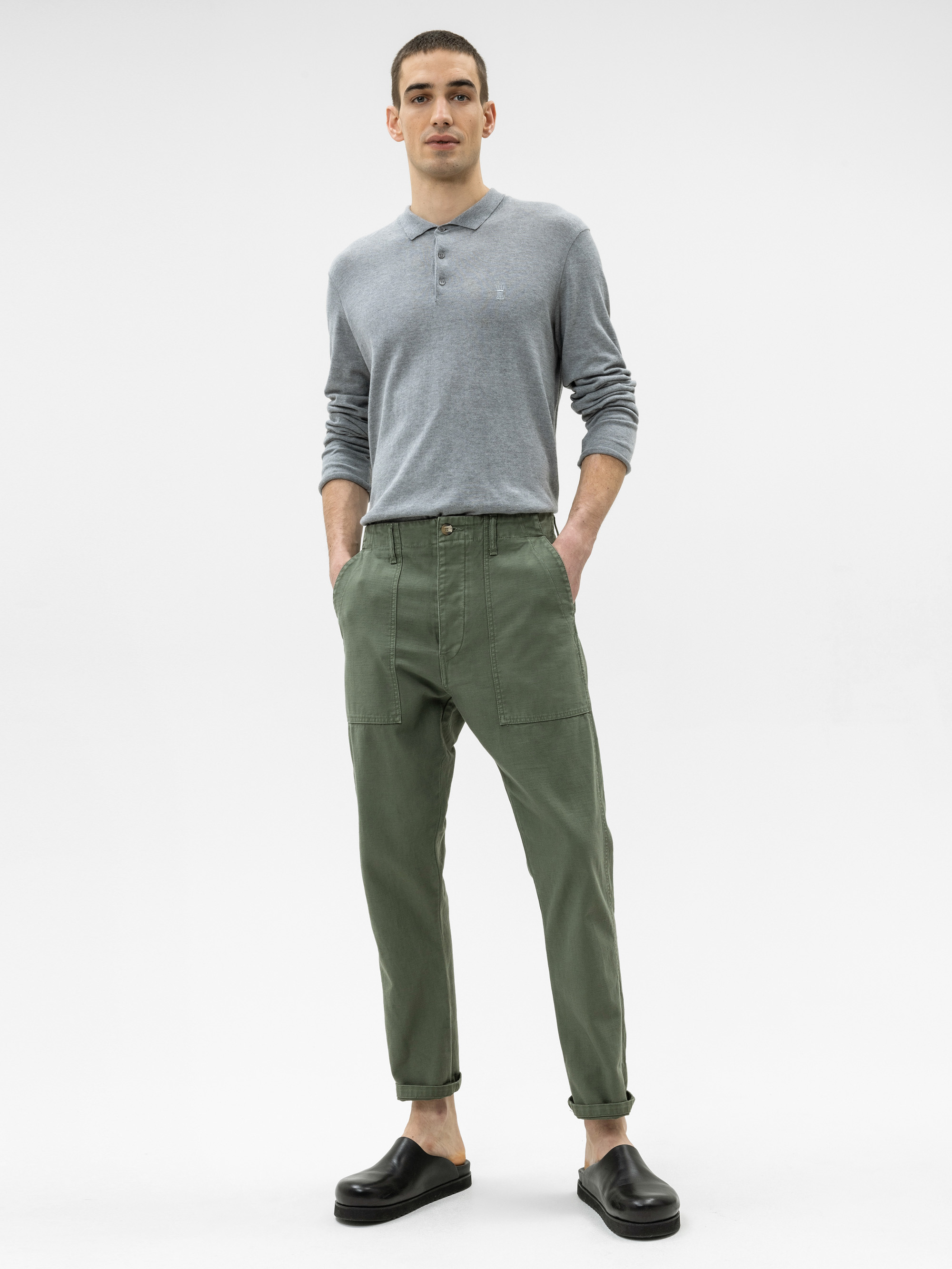 Eine Person steht vor einem weißen Hintergrund und trägt ein hellgraues, langärmeliges Henley-Shirt, eine olivgrüne Hose mit großen Vordertaschen und schwarze Slipper. Die Hände befinden sich in den Taschen und der Blick ist nach vorne gerichtet.