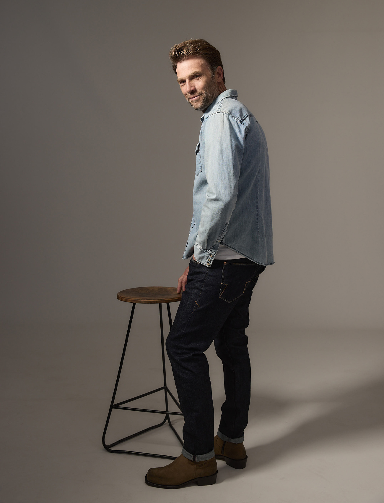 Ein Mann in einem hellen Jeanshemd und dunklen Jeans steht neben einem hohen Holzhocker, schaut über die Schulter und lächelt, in einem Studio mit neutralem Hintergrund.
