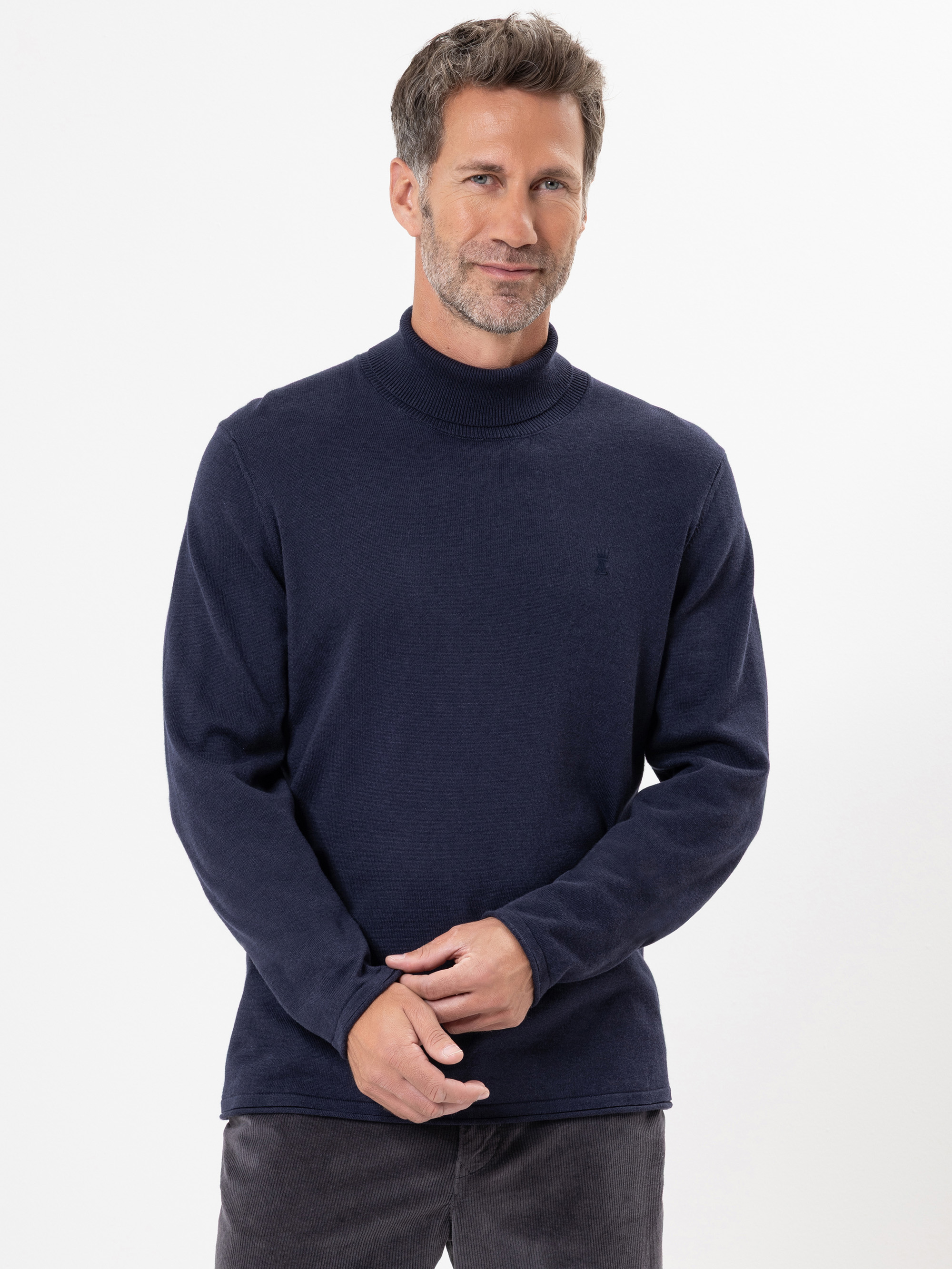 Ein Mann mit kurzen braunen Haaren und Bart steht vor einem weißen Hintergrund, trägt einen marineblauen Rollkragenpullover und eine dunkle Hose und hat die Hände leicht vor sich verschränkt.