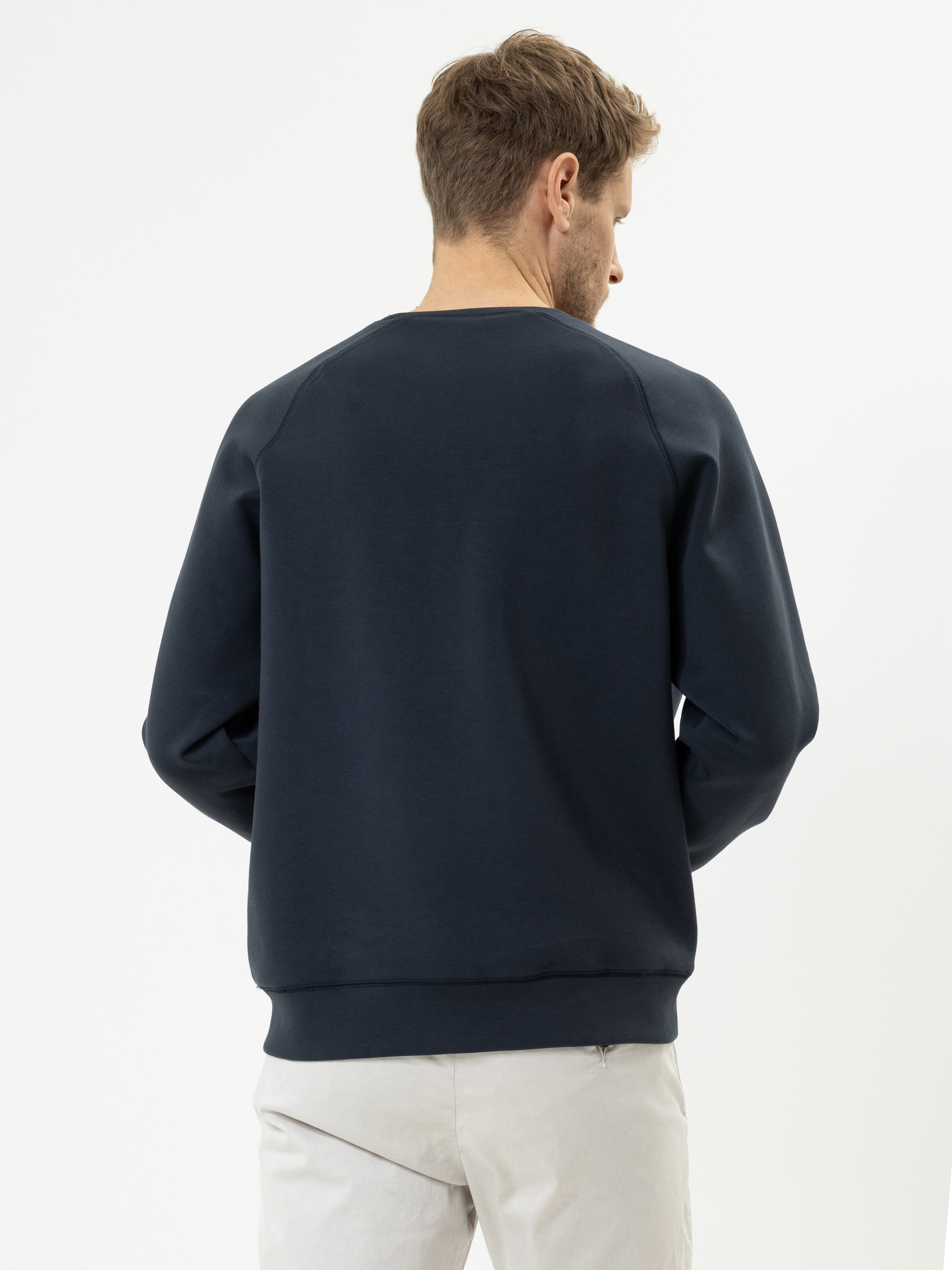 Ein Mann mit hellbraunem Haar ist von hinten zu sehen. Er trägt ein dunkles, marineblaues Sweatshirt und eine helle Hose und steht vor einem schlichten weißen Hintergrund.