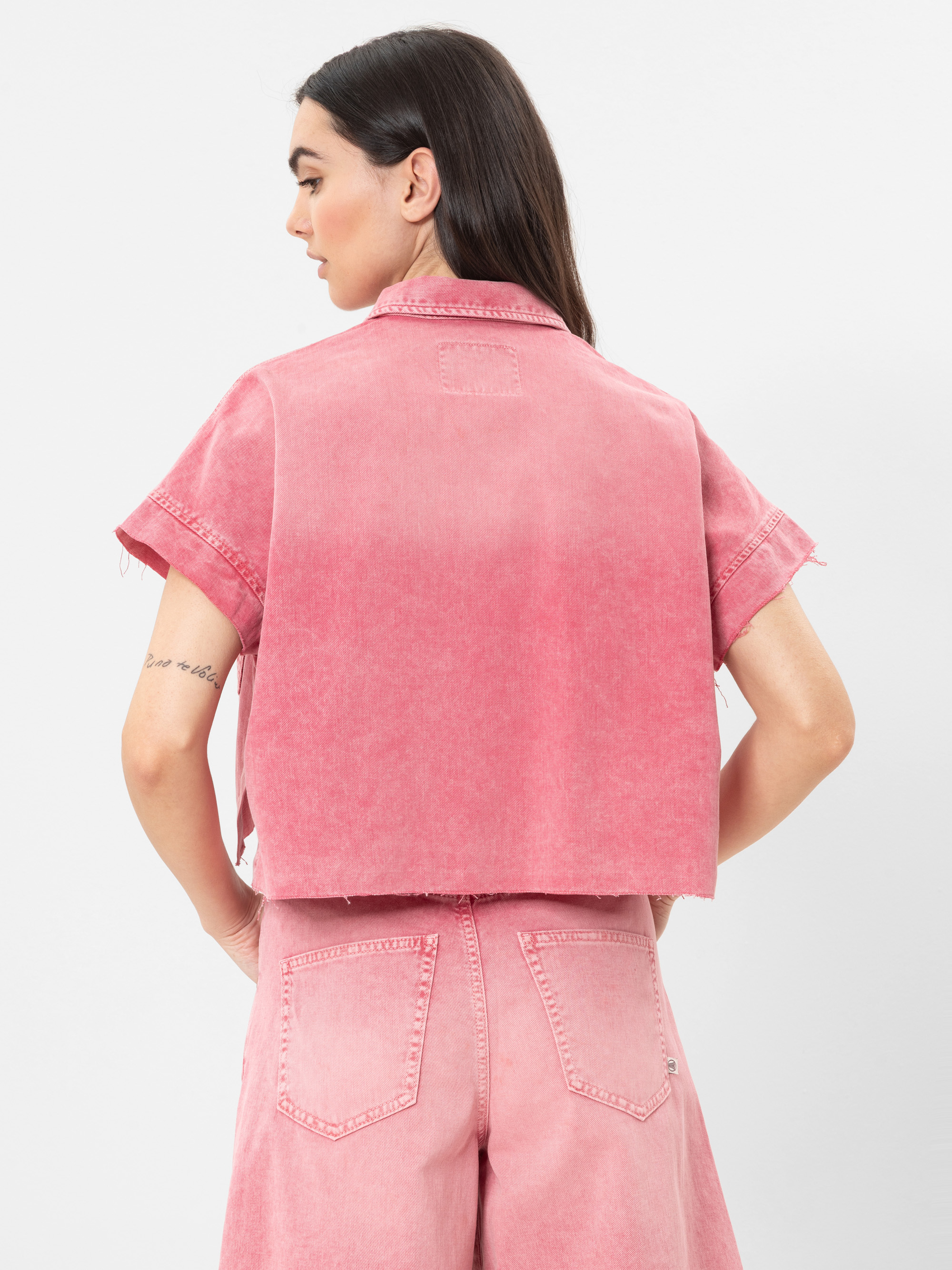 Eine Frau mit langen dunklen Haaren ist von hinten zu sehen. Sie trägt eine kurzärmelige pinkfarbene Jeansjacke und dazu passende hochgeschnittene pinkfarbene Jeansshorts. Sie hat eine Tätowierung auf ihrem linken Arm und steht vor einem unifarbenen Hi