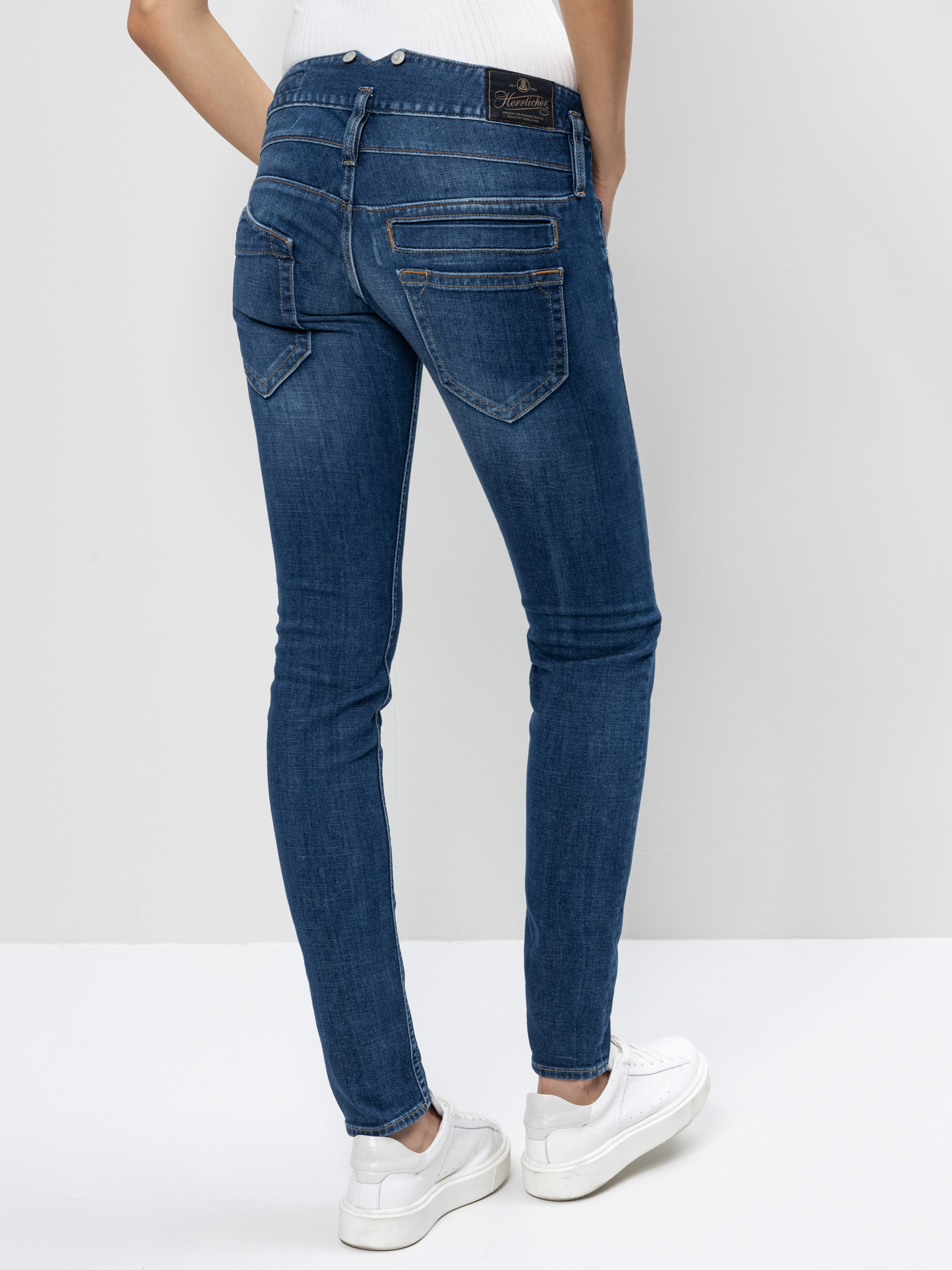 Eine Person in blauen Skinny-Jeans und weißen Turnschuhen steht mit dem Gesicht zur Kamera vor einem schlichten, hellen Hintergrund.