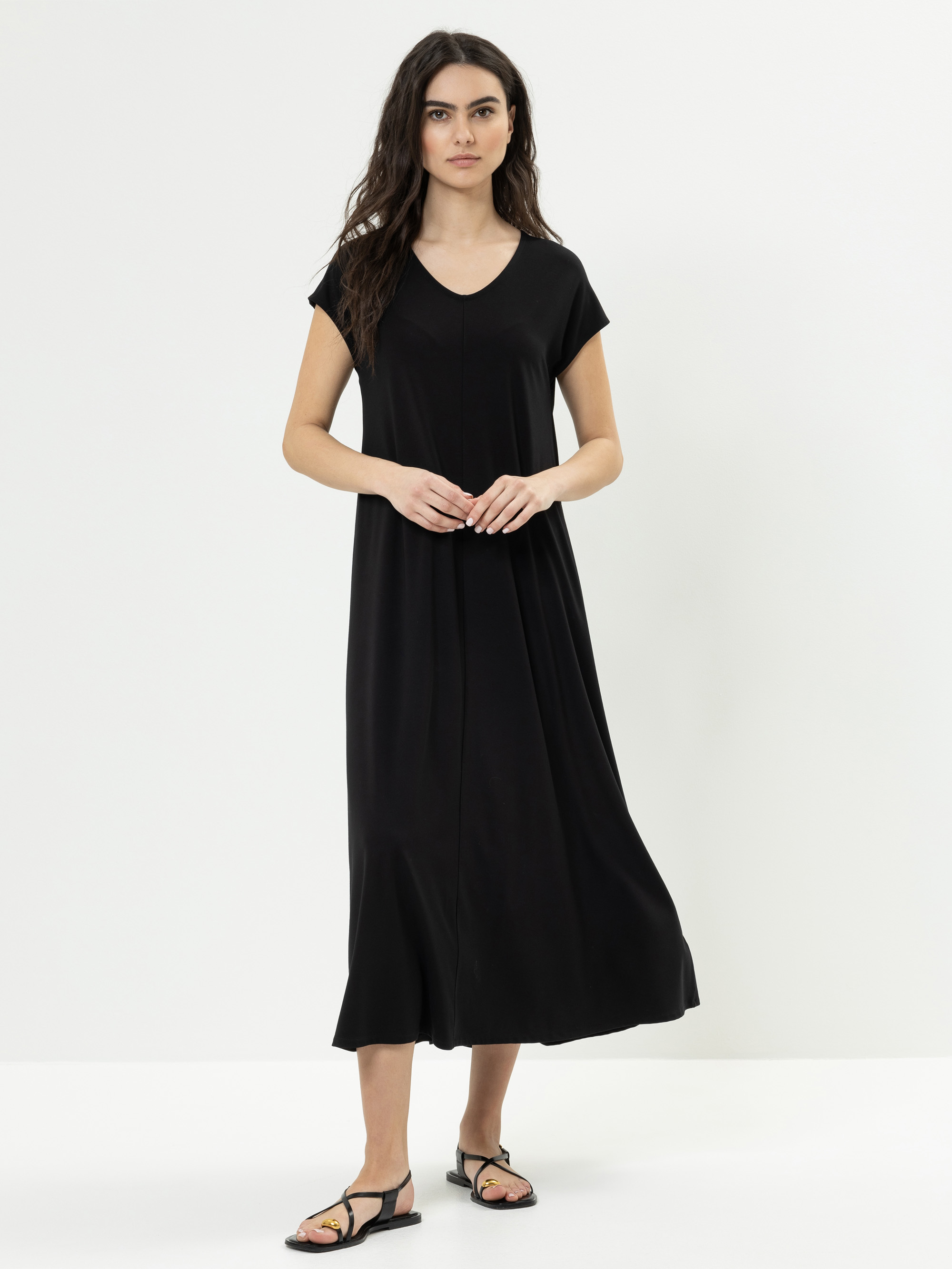Eine Frau mit langen dunklen Haaren trägt ein locker sitzendes, kurzärmeliges schwarzes Midikleid mit V-Ausschnitt und schwarze Sandalen und steht vor einem schlichten weißen Hintergrund.