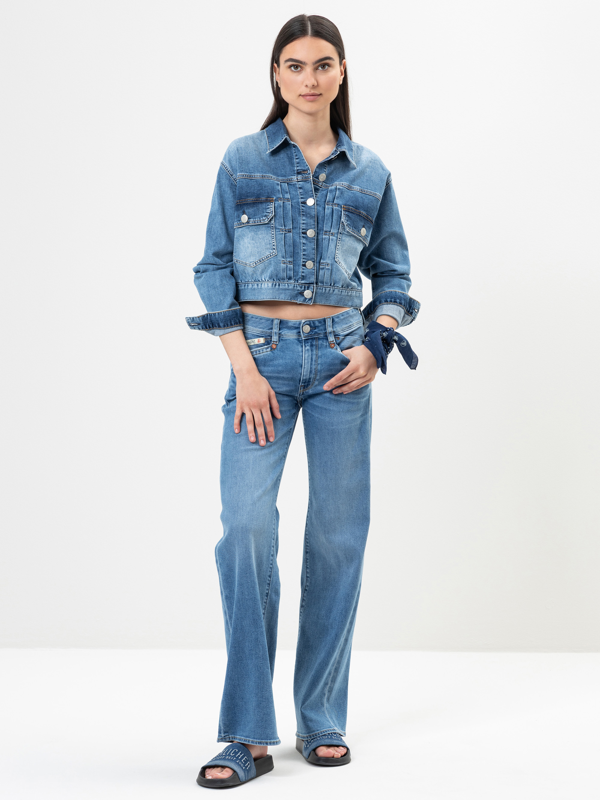 Eine Frau steht vor einem einfarbigen Hintergrund und trägt eine abgeschnittene Jeansjacke, hochgekrempelte Bluejeans, schwarze Slide-Sandalen und ein blaues Halstuch, das sie um ihr linkes Handgelenk gebunden hat. Sie blickt mit neutralem Blick nach vor