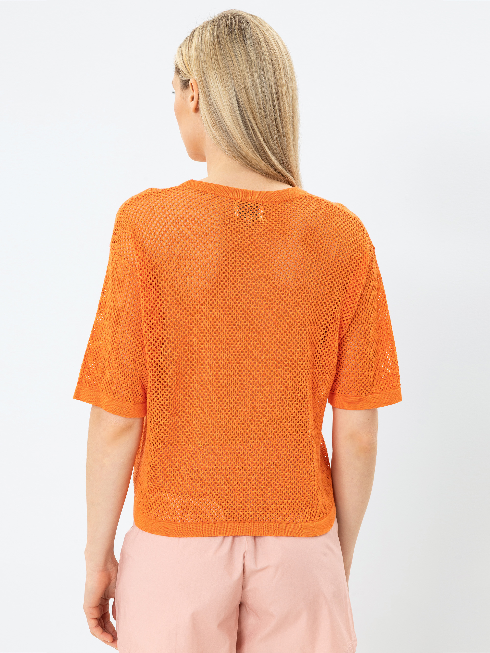Eine Person mit langen blonden Haaren ist von hinten zu sehen. Sie trägt ein orangefarbenes kurzärmeliges Netzoberteil und hellrosa Shorts vor einem schlichten weißen Hintergrund.