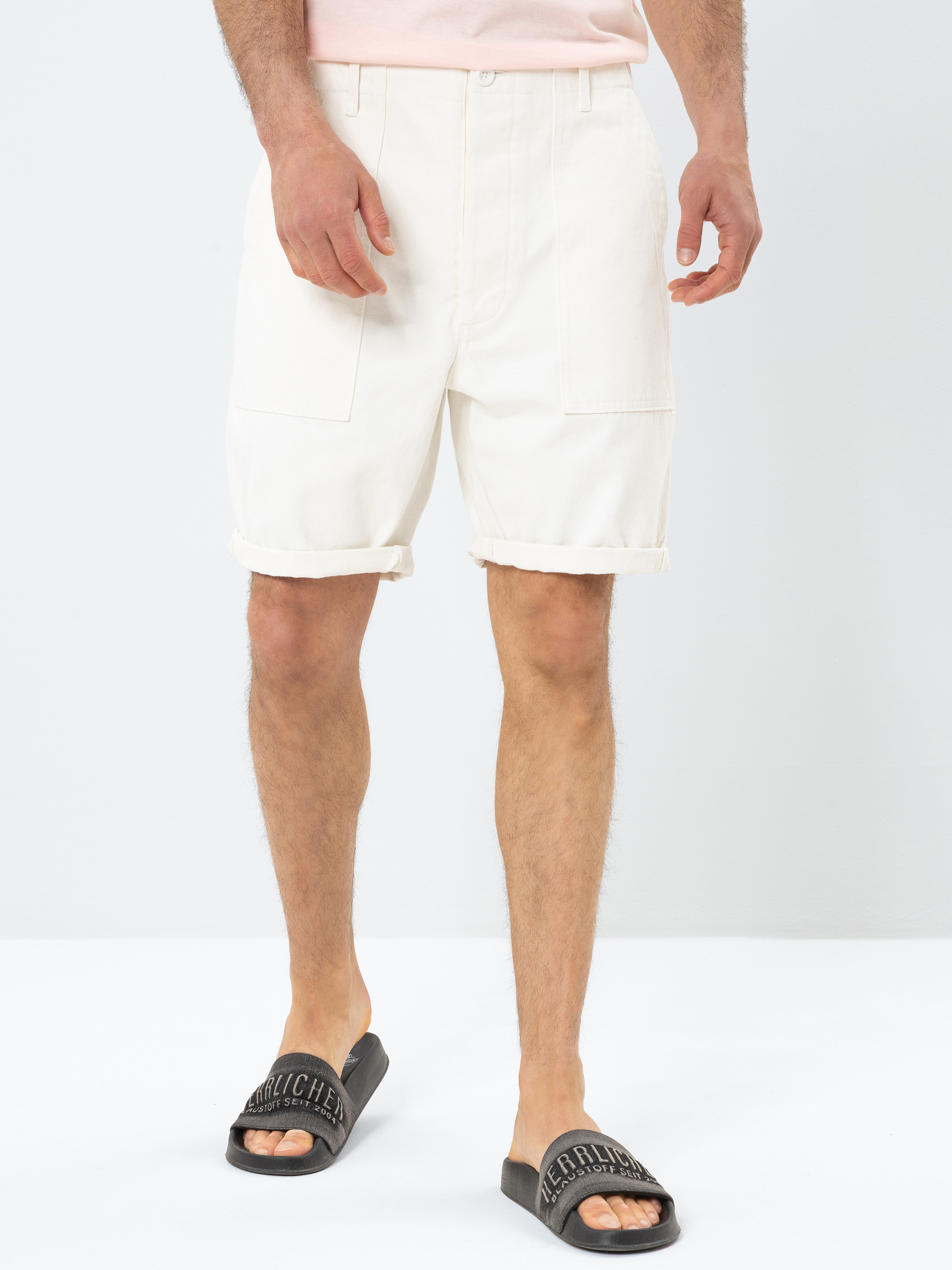Eine Person steht vor einem einfarbigen Hintergrund und trägt knielange, weiße Shorts, ein hellrosa Hemd und schwarze Slide-Sandalen. Nur die untere Hälfte ihres Körpers ist sichtbar.