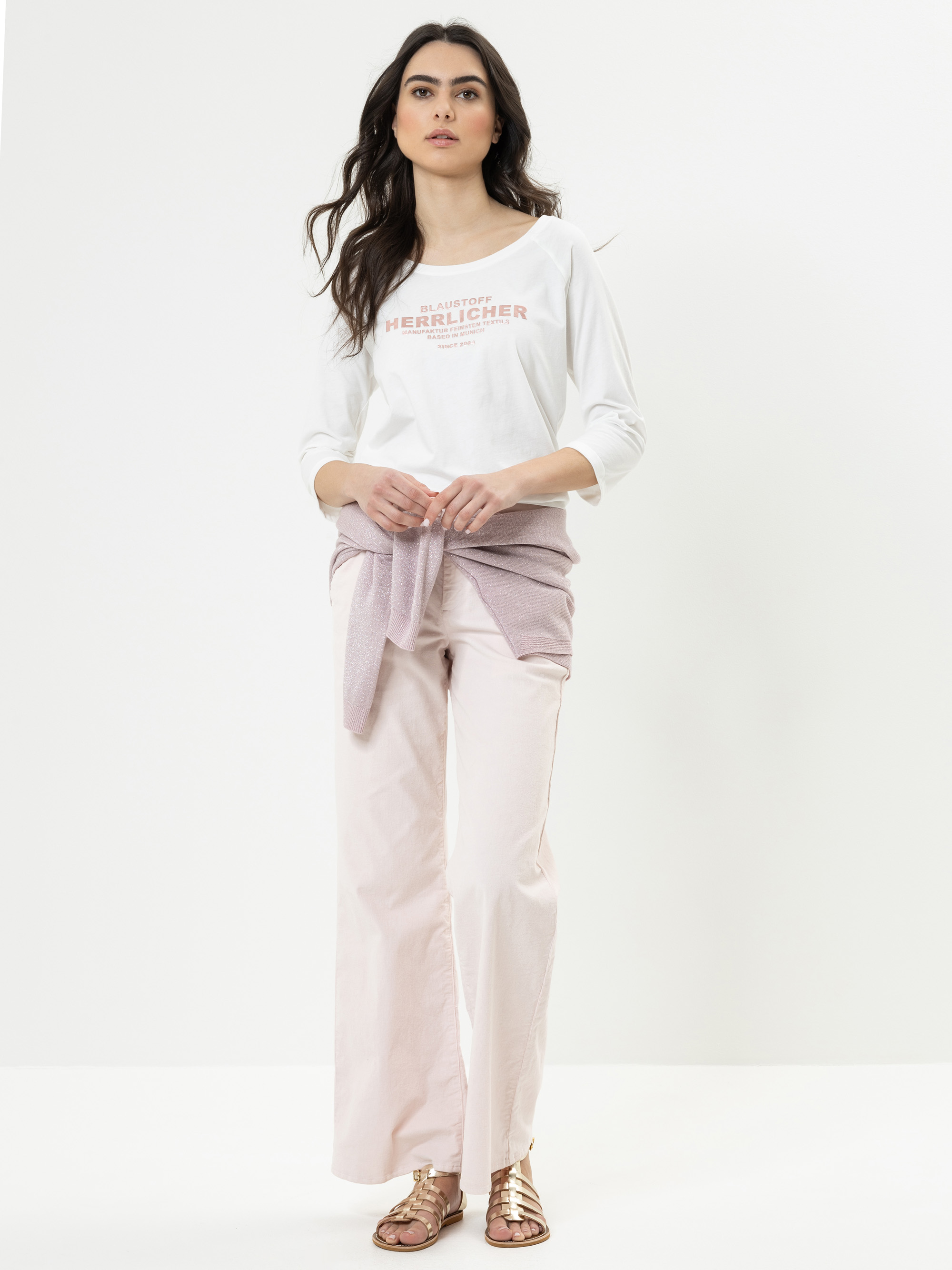 Eine Frau mit langen dunklen Haaren steht vor einem schlichten weißen Hintergrund. Sie trägt ein weißes langärmeliges Oberteil mit Text, eine hellrosa Hose mit weitem Bein, einen hellvioletten Pullover, den sie um die Taille bindet, und metallische Sa