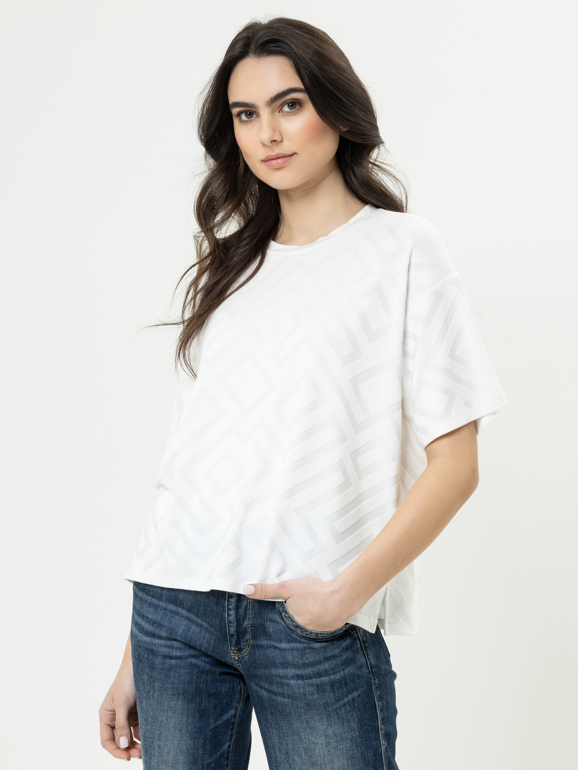 Eine Frau mit langen braunen Haaren trägt ein lockeres, weißes T-Shirt mit geometrischem Muster und blaue Jeans. Sie steht vor einem schlichten weißen Hintergrund und hat eine Hand in der Tasche.