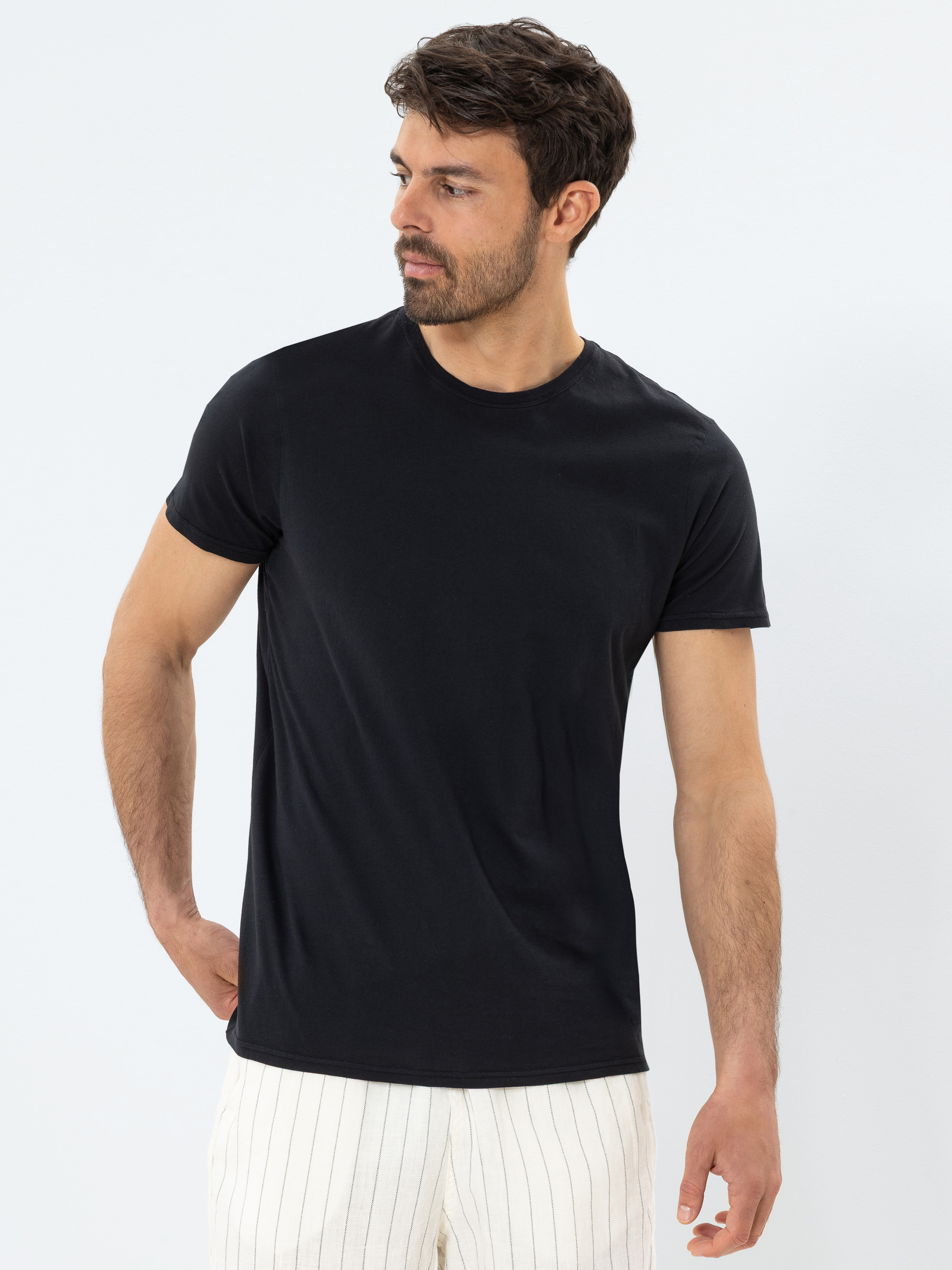 Ein Mann mit kurzen dunklen Haaren und einem Bart trägt ein schlichtes schwarzes T-Shirt und eine weiße Hose mit dünnen vertikalen Streifen. Er steht vor einem hellen Hintergrund und blickt nach links, eine Hand hinter dem Rücken.