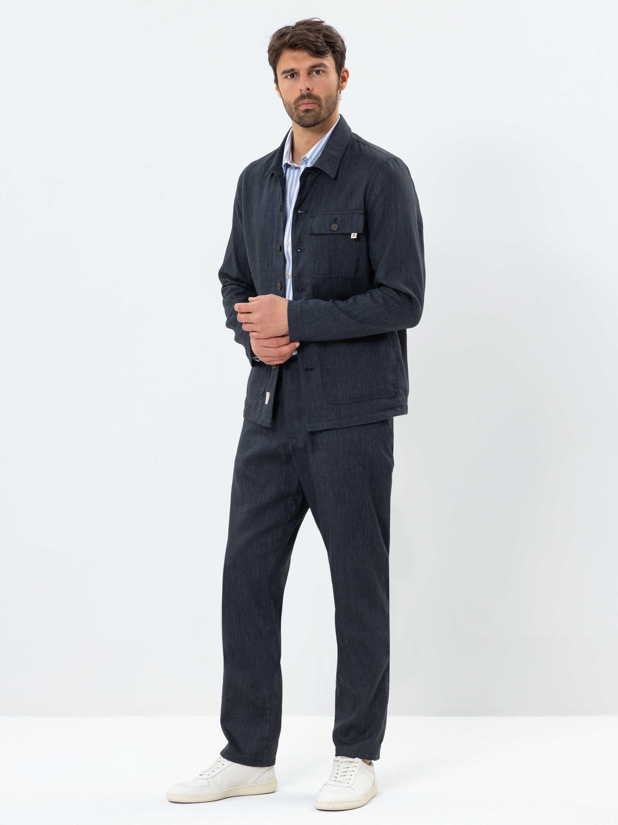 Ein Mann steht vor einem schlichten weißen Hintergrund und trägt eine dunkelblaue Jacke und eine passende Hose, ein hellblau gestreiftes Hemd und weiße Turnschuhe. Er hat kurzes dunkles Haar und einen Bart und blickt direkt in die Kamera.