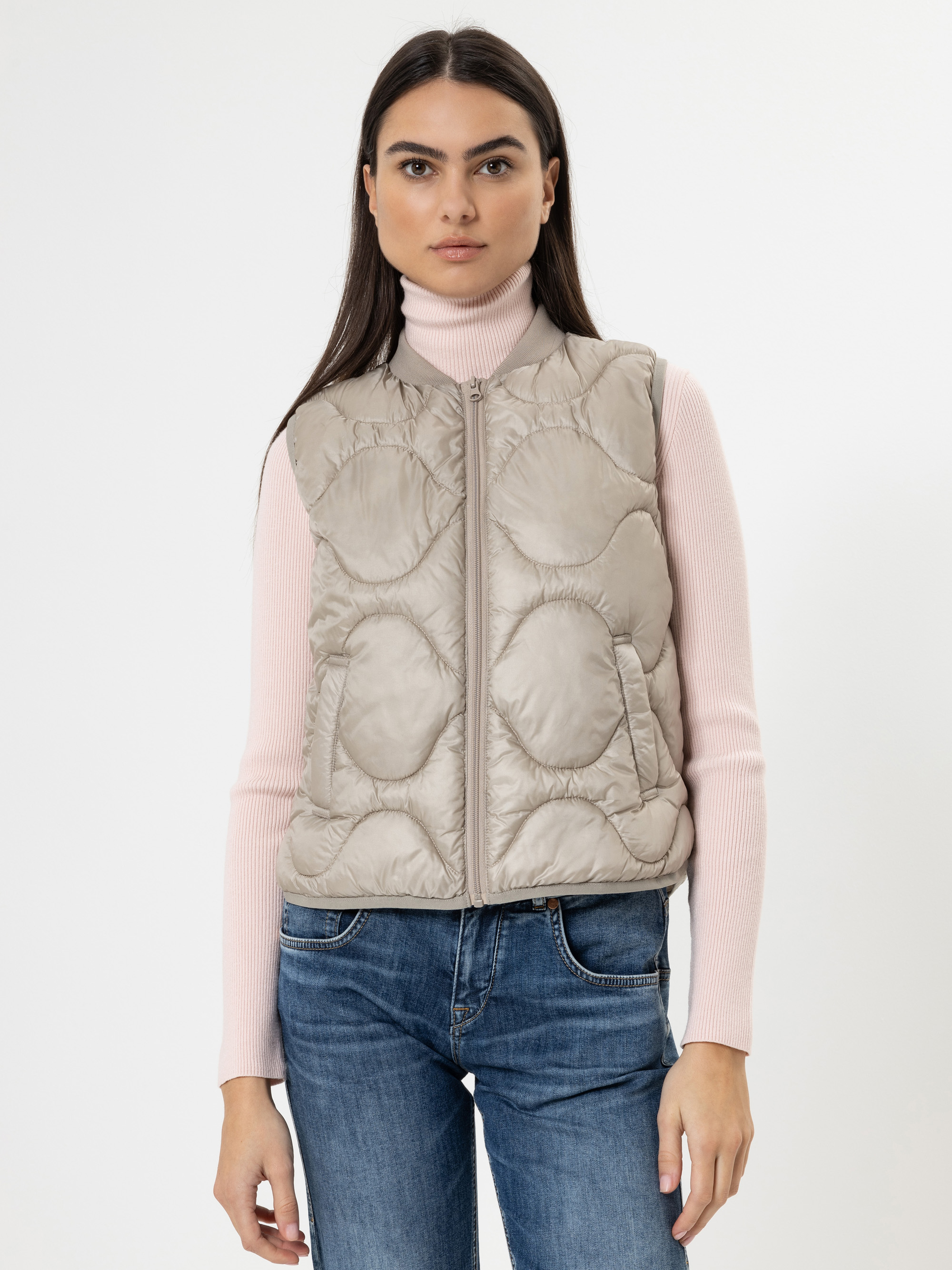 Eine Frau mit langen dunklen Haaren trägt einen hellrosa Rollkragenpullover, eine beige Steppweste und blaue Jeans. Sie steht mit dem Gesicht nach vorne vor einem schlichten weißen Hintergrund.