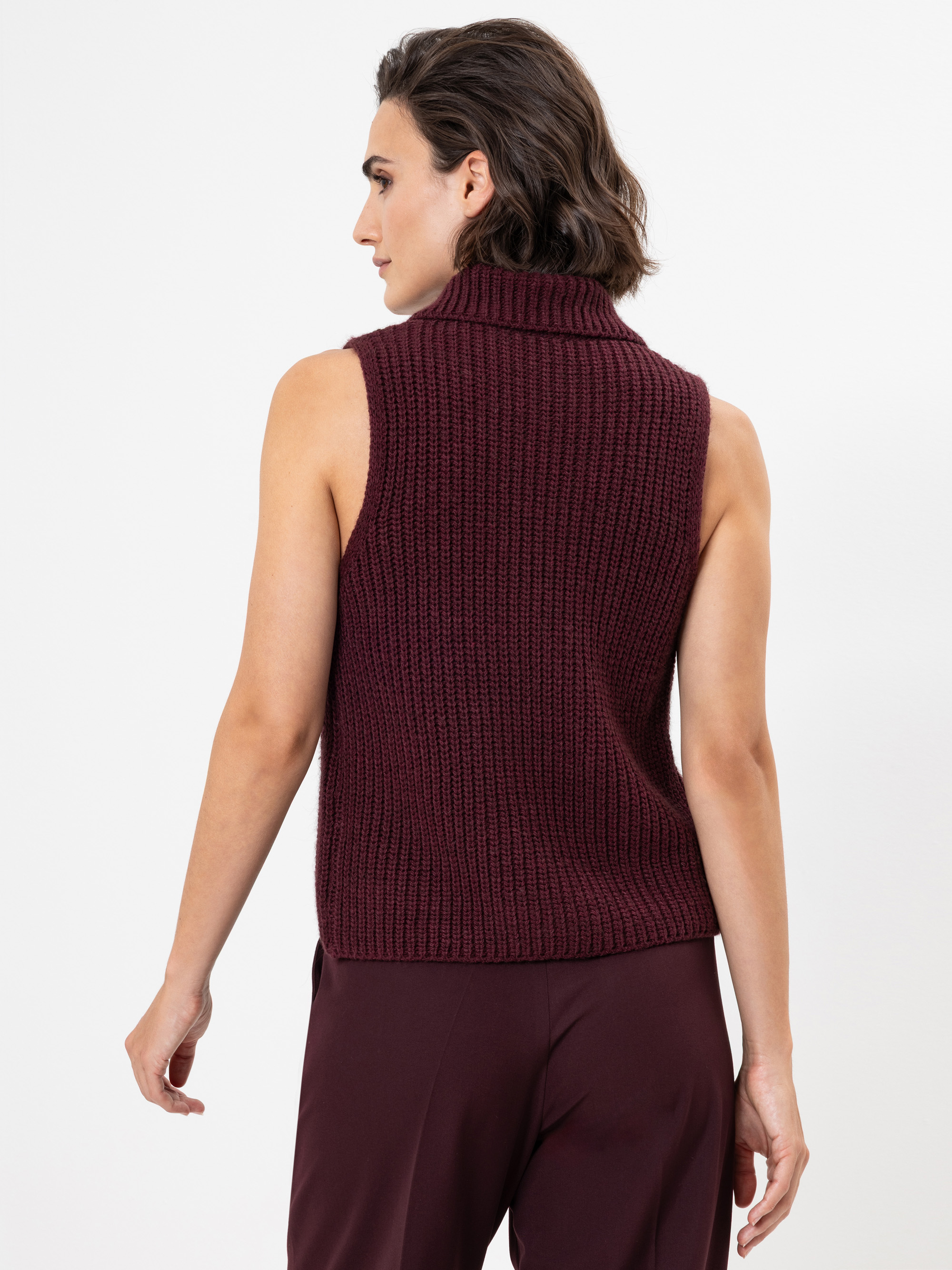 Eine Person mit kurzen braunen Haaren wird von hinten gezeigt. Sie trägt einen ärmellosen, dunkelburgunderfarbenen Rollkragenpullover und eine passende burgunderfarbene Hose vor einem schlichten weißen Hintergrund.