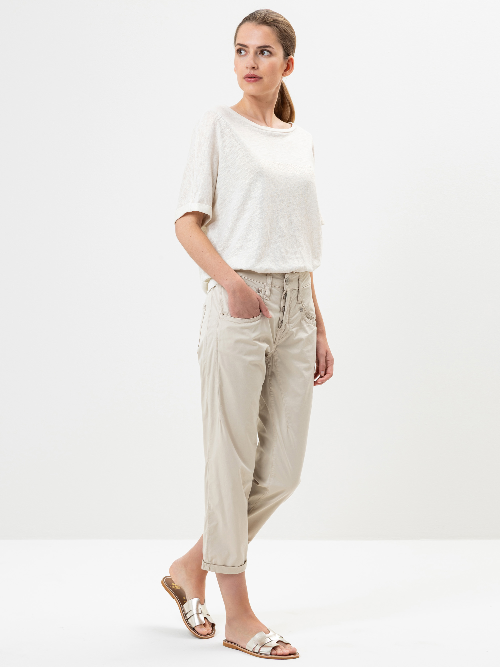 Eine Frau steht vor einem einfarbig weißen Hintergrund. Sie trägt ein lockeres weißes kurzärmeliges Oberteil, eine hellbeige Hose mit aufgerollten Manschetten und metallische Sandalen. Sie hat die Hände in den Taschen und blickt zur Seite.